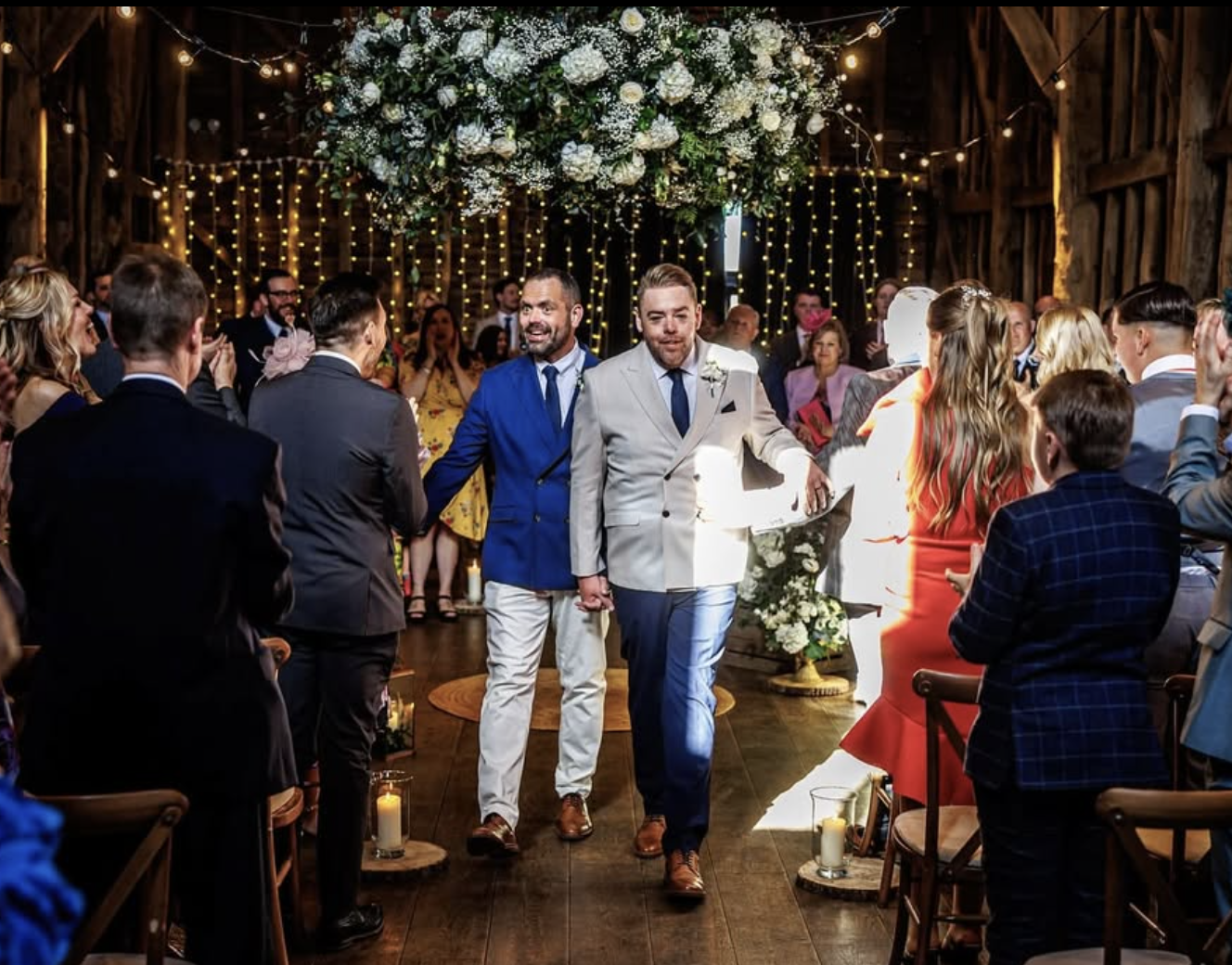 Two men in suits holding hands, walking down the aisle at a wedding ceremony inside a rustic barn decorated with string lights and a large floral arrangement overhead, surrounded by guests clapping.