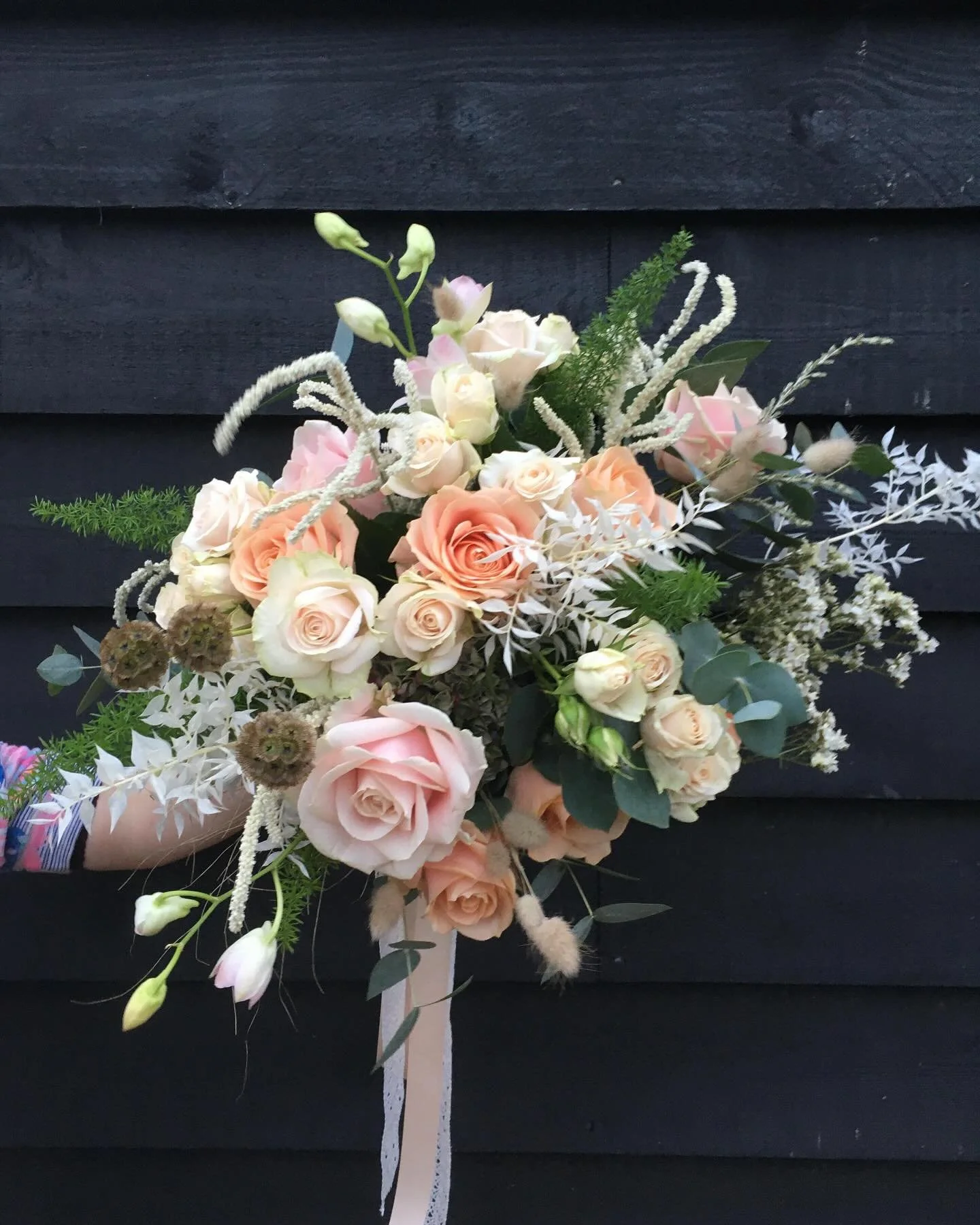 A bouquet of pale pink and white roses, white and pink lilies, green leaves, and decorative white, fuzzy, curly stems against a dark background.