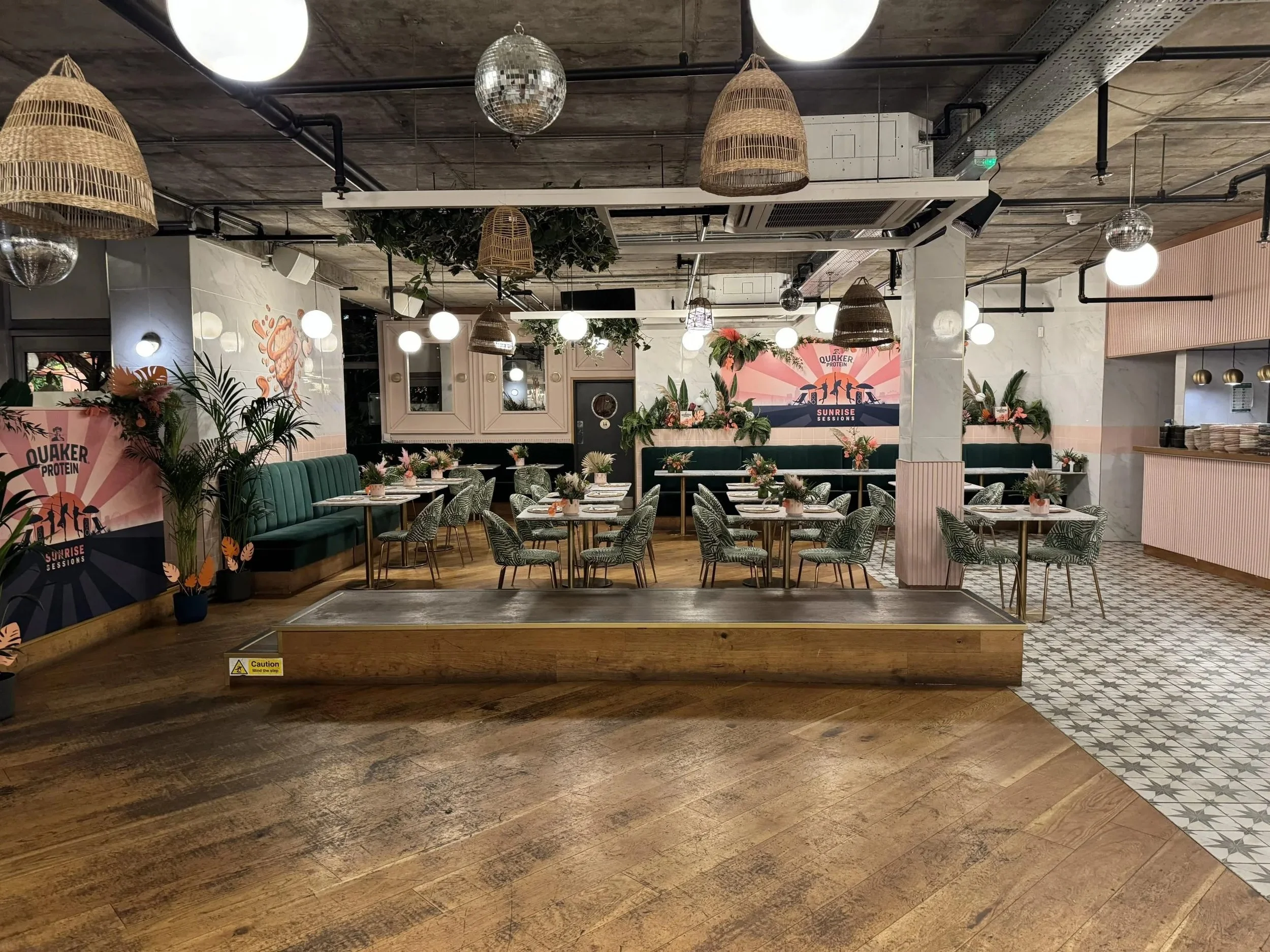Decorated dining area with tables, chairs, potted plants, and floral arrangements in a modern restaurant with patterned tiles and wooden flooring.