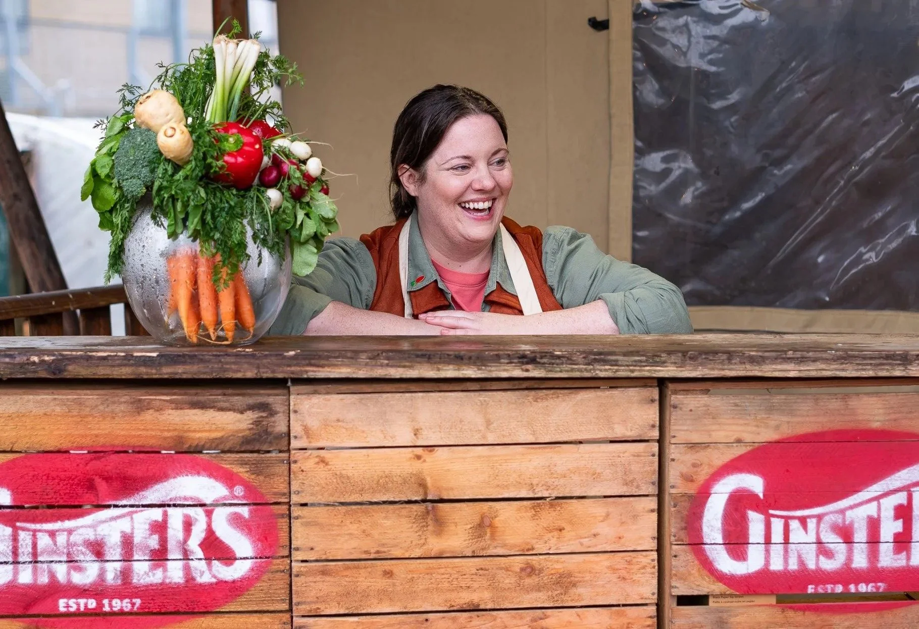 Woman smiling behind a wooden bar with a large basket of fresh vegetables, including carrots, radishes, celery, and a red bell pepper, in front of a Ginsters sign.