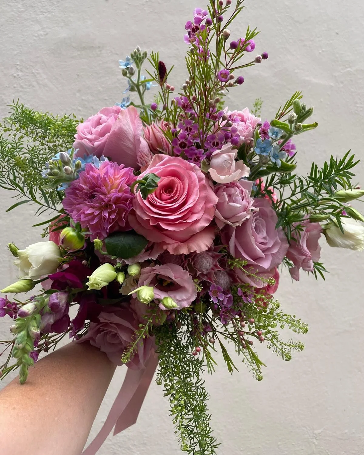 A colorful bouquet of pink, white, purple, and blue flowers with green foliage, held by a person's arm against a neutral background.