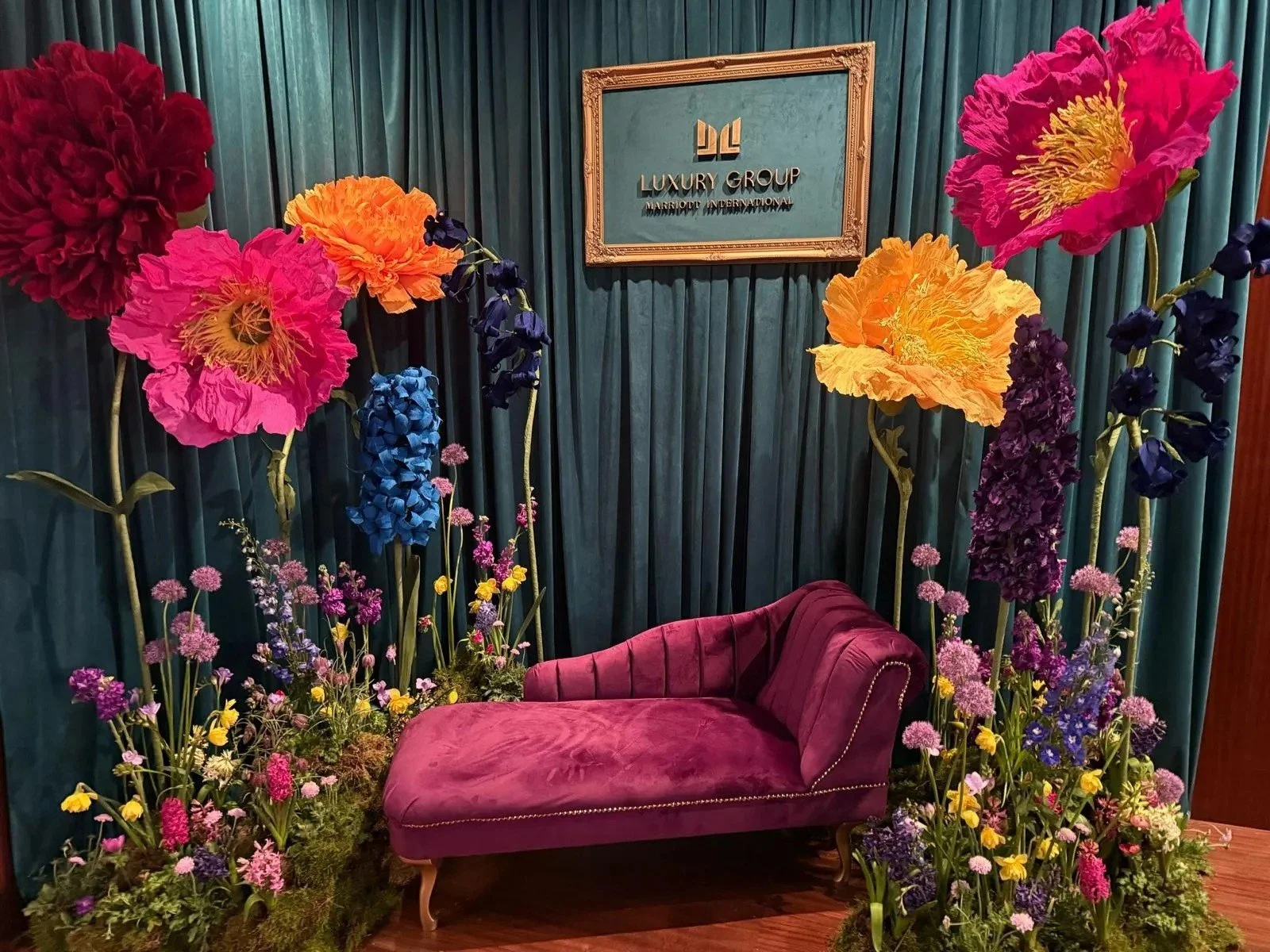 A luxurious interior setting with a pink velvet chaise lounge surrounded by large colorful paper flowers and and a sign that reads 'LUXURY GROUP' on the wall.