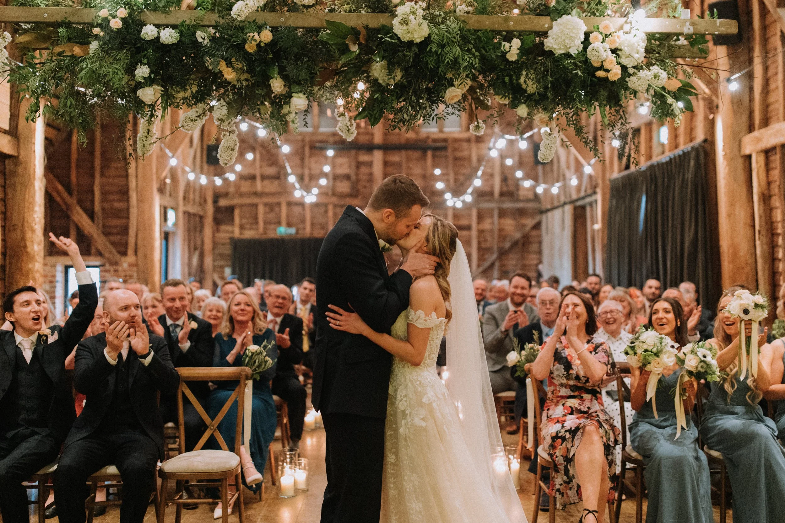 A couple shares a kiss during their wedding ceremony in a rustic barn, surrounded by seated guests clapping and smiling, with floral decorations and string lights overhead.
