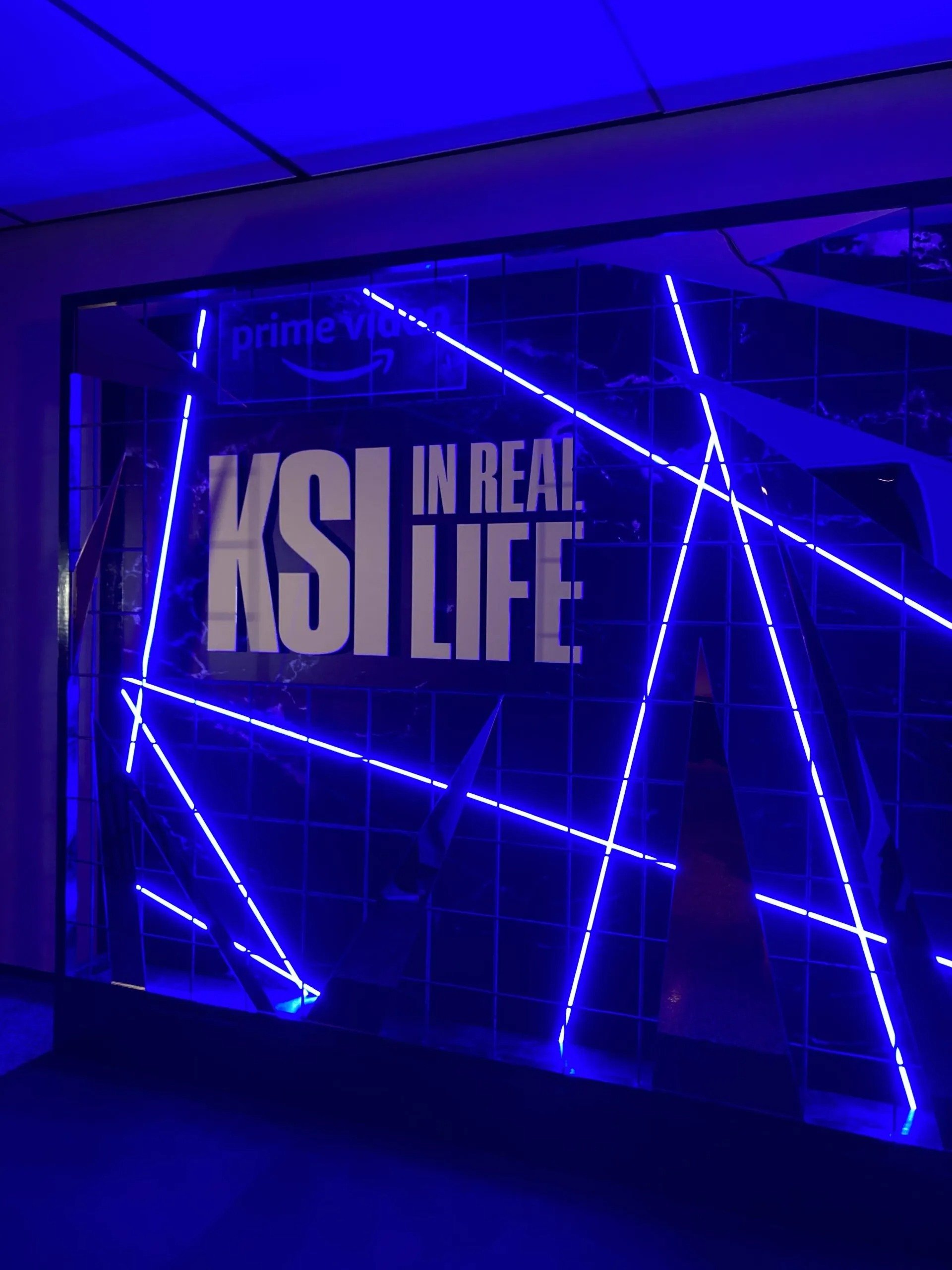 Neon sign with blue lights and white text reading 'KSI IN REAL LIFE' behind a grid of glass panels, with a small Amazon Prime Video logo in the top left corner.