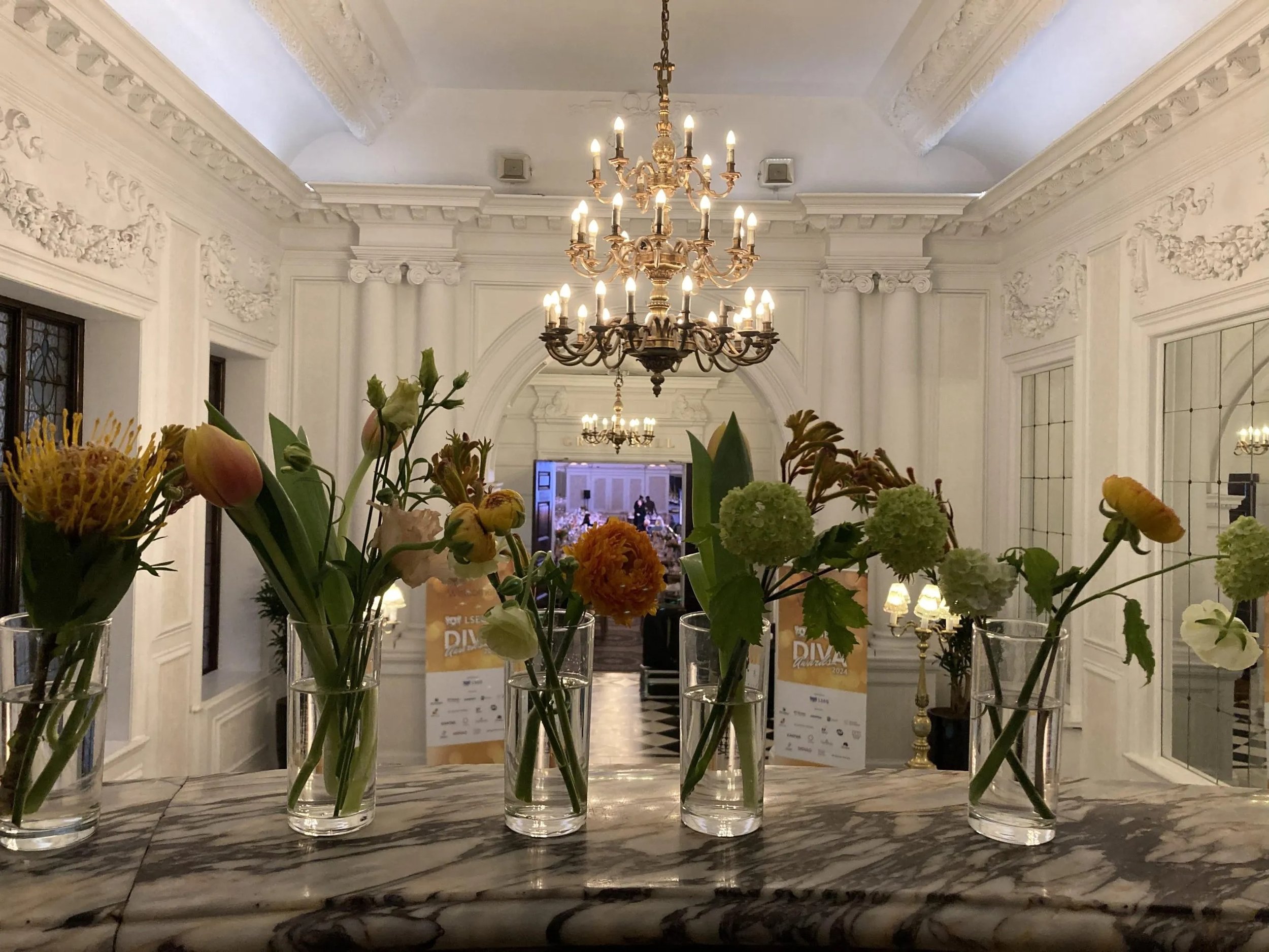 Elegant hall with white ornate walls, chandeliers, and a table with attached vases holding various flowers.