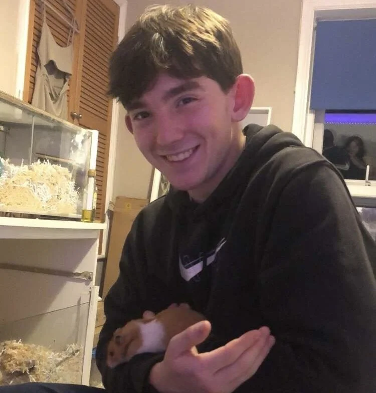 A young man with short dark hair smiling at the camera, holding a small hamster in his hands. In the background, there is an enclosure with bedding and a few items inside, and a window showing a purple-lit room.