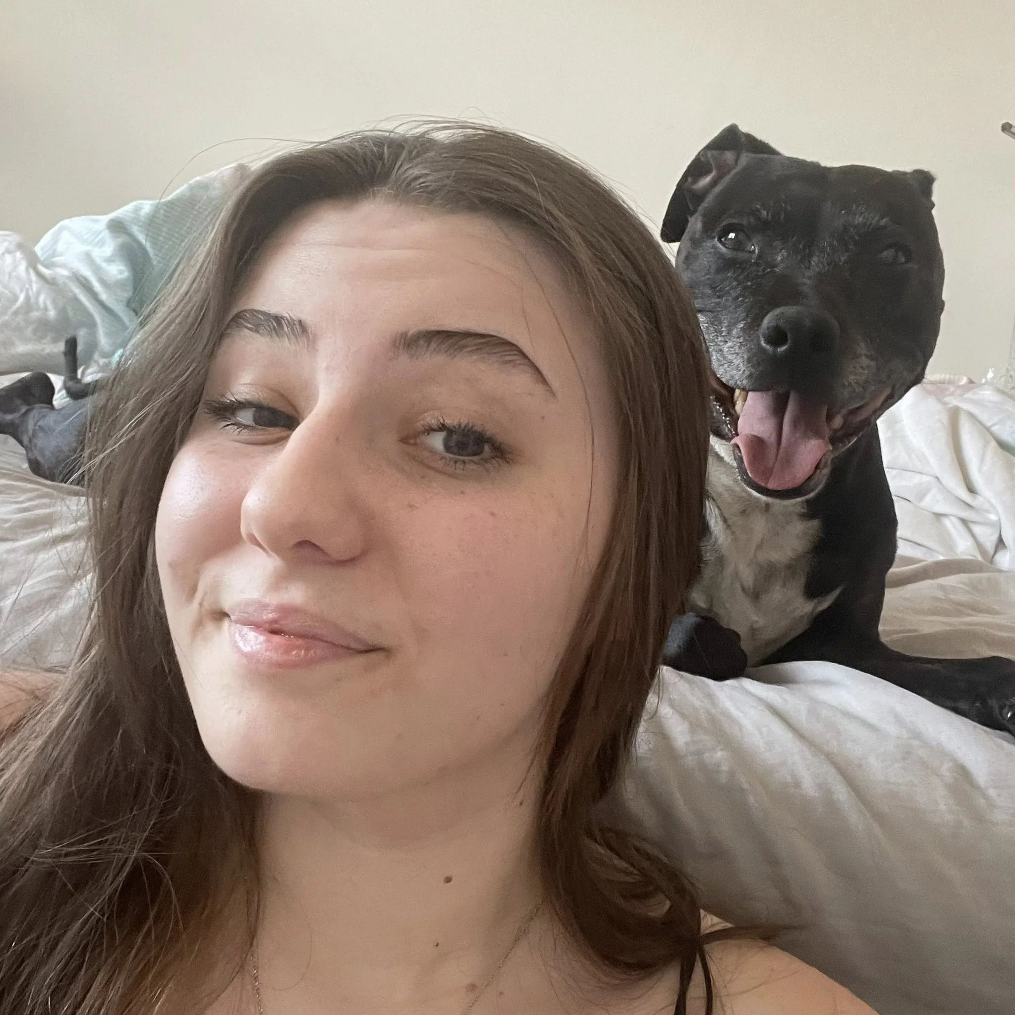 A young woman with brown hair taking a selfie with a smiling black and white dog behind her on a bed.