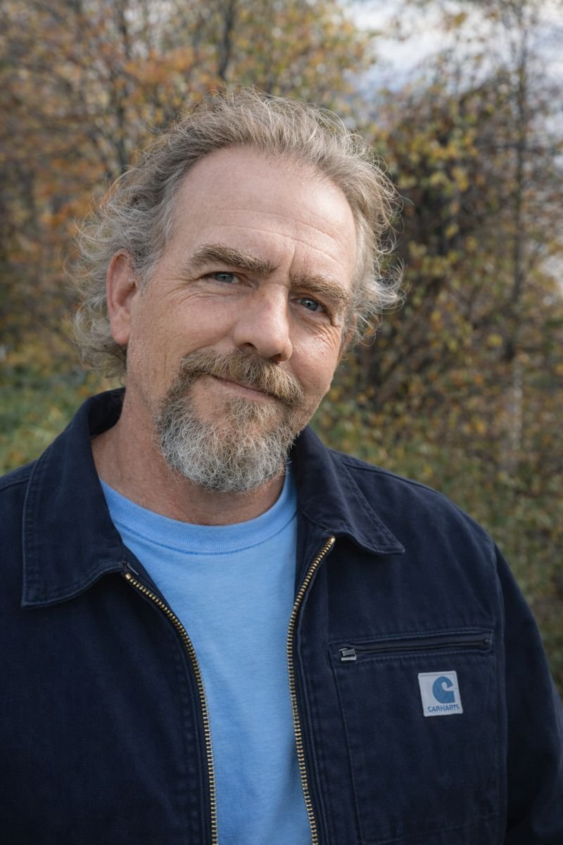 A middle-aged man with gray hair, a beard, and blue eyes smiling outdoors with autumn trees in the background, wearing a black jacket and a light blue shirt.