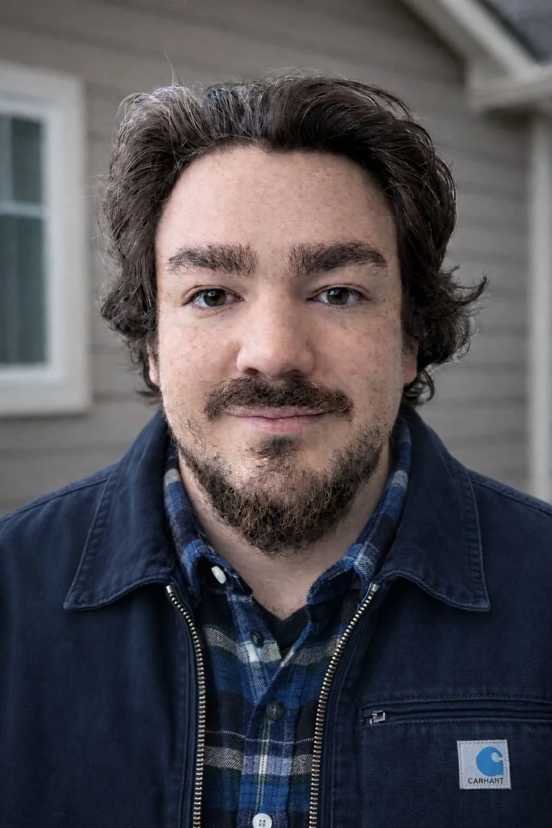 A close-up portrait of a man with wavy dark hair, light skin, and a beard, wearing a blue plaid shirt and a navy Carhartt jacket, standing outside in front of a wooden house with a window.
