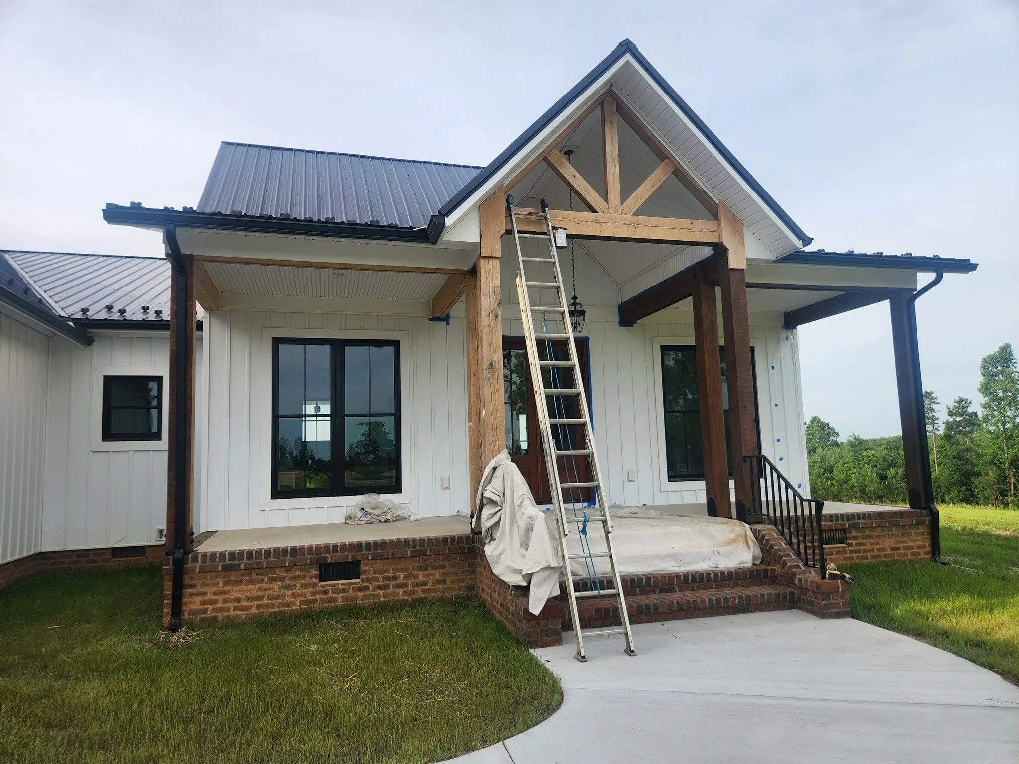House under construction with a front porch, ladder, and partially completed wooden beams, surrounded by green grass and trees in the background.
