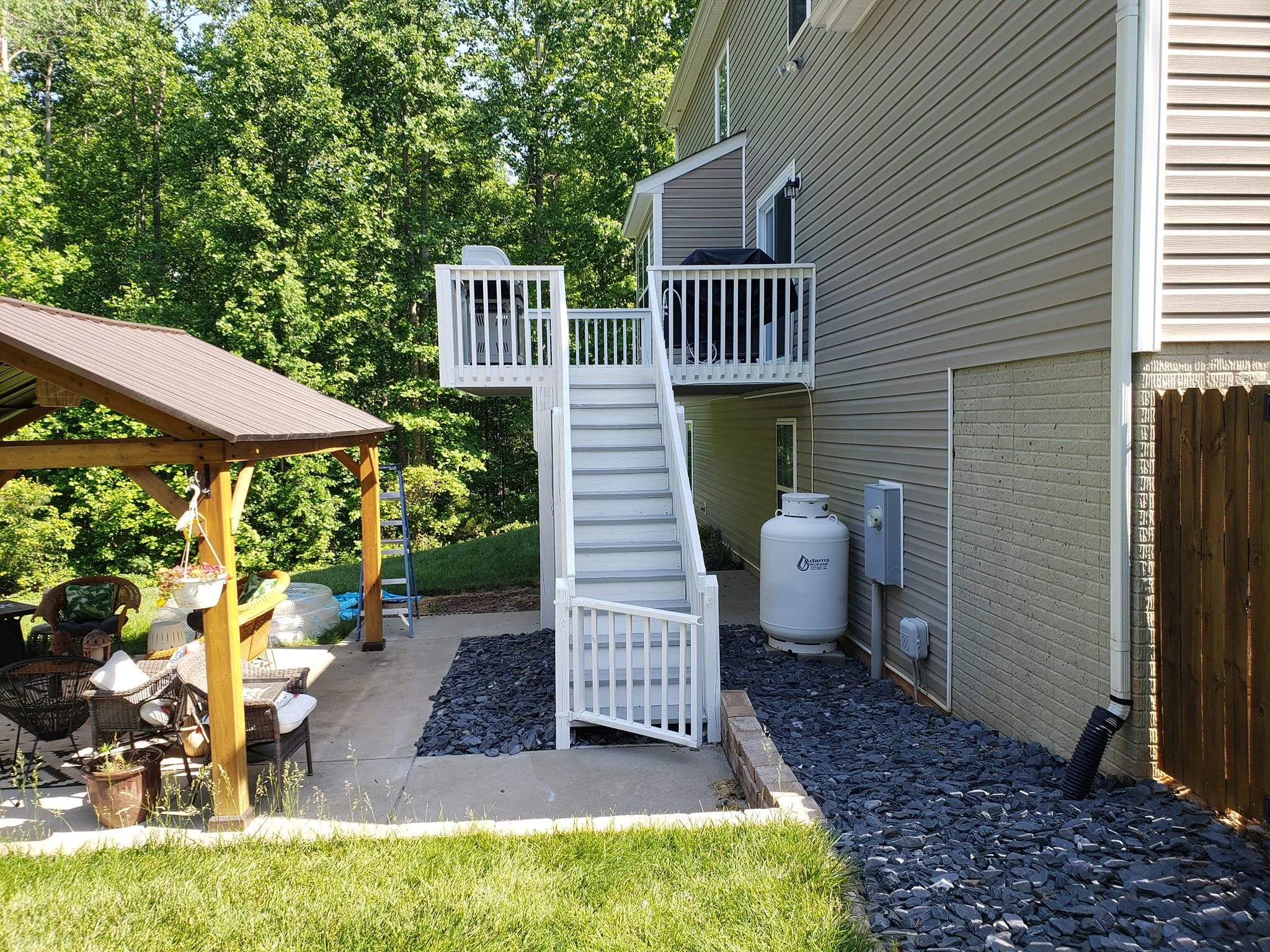 Backyard with a beige house, a white staircase leading to a second-story deck, a wooden shed with outdoor furniture, and trees in the background.