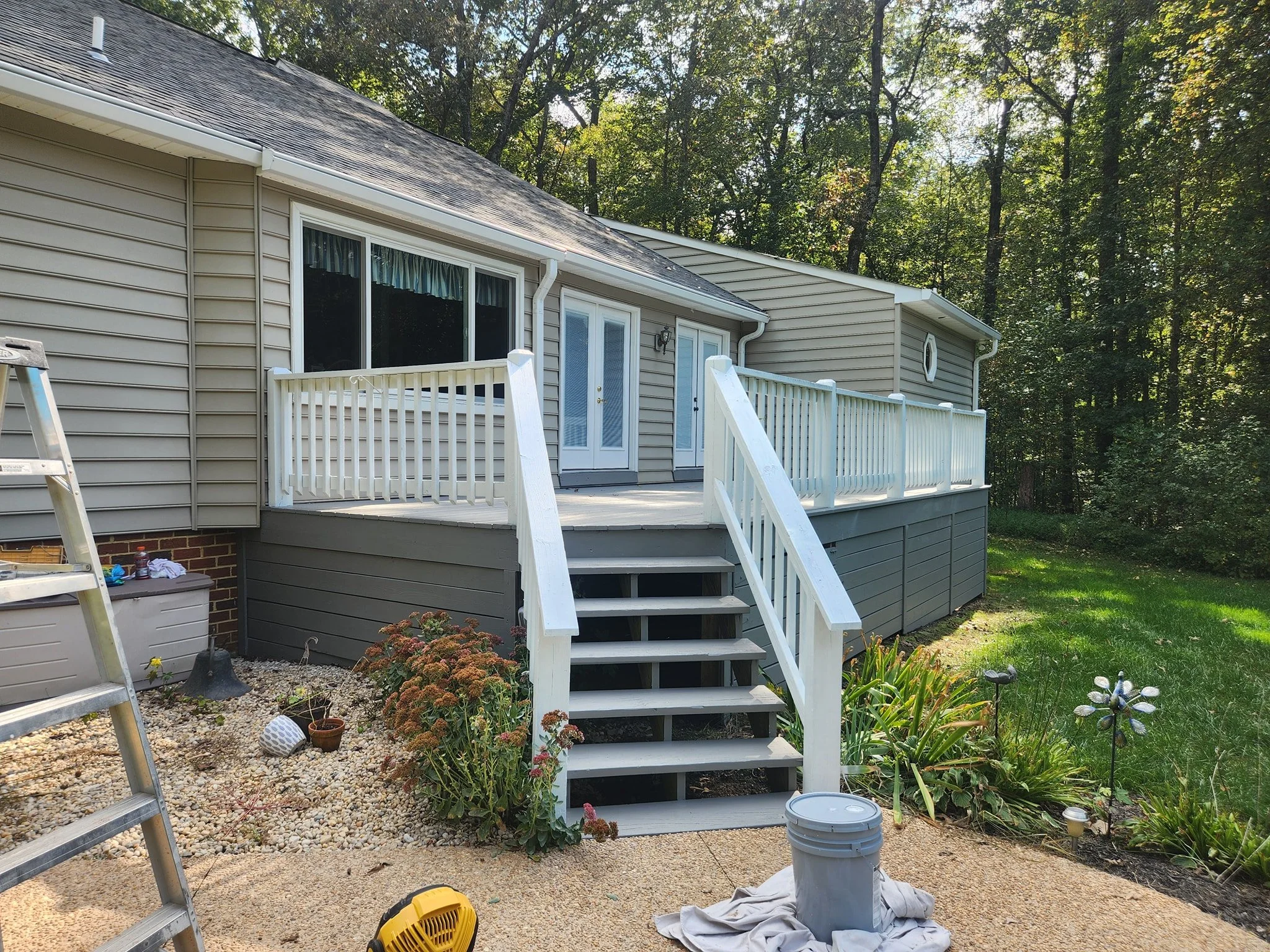A backyard deck attached to a house, with stairs leading down to a garden area with plants, flowers, and yard lights. There are tools and a bucket nearby, indicating ongoing outdoor work.
