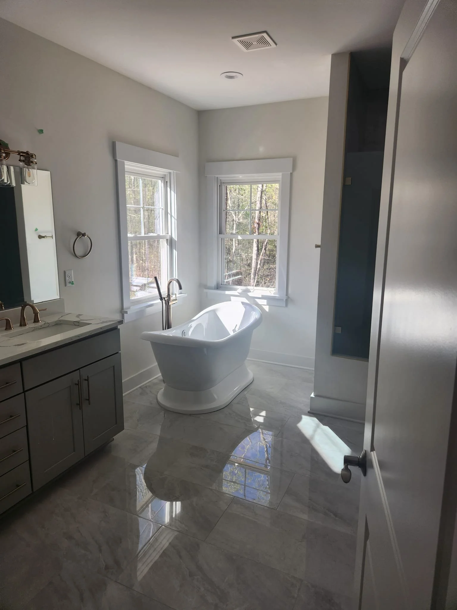 Bright bathroom with two windows, a white bathtub, grey vanity with a marble countertop, and tiled flooring.