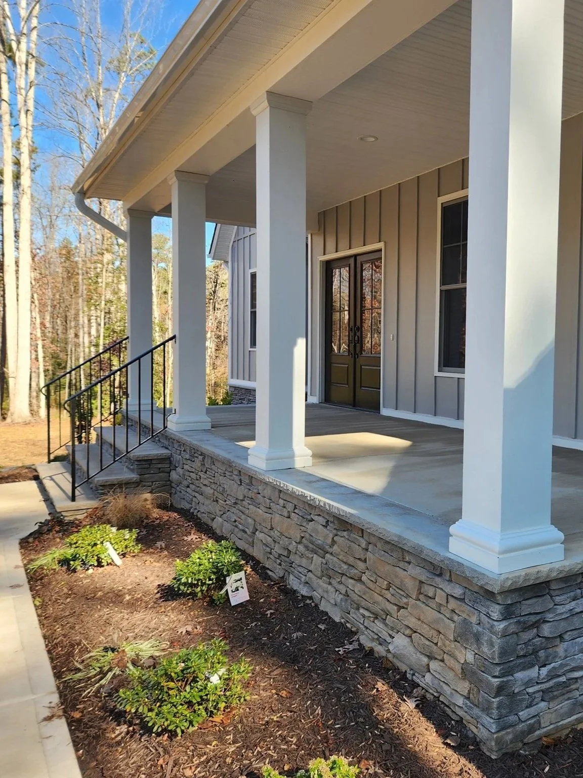 Front porch of a house with four white columns, a stone foundation, and black railing, with small landscaped bushes beneath, and a wooded area in the background.