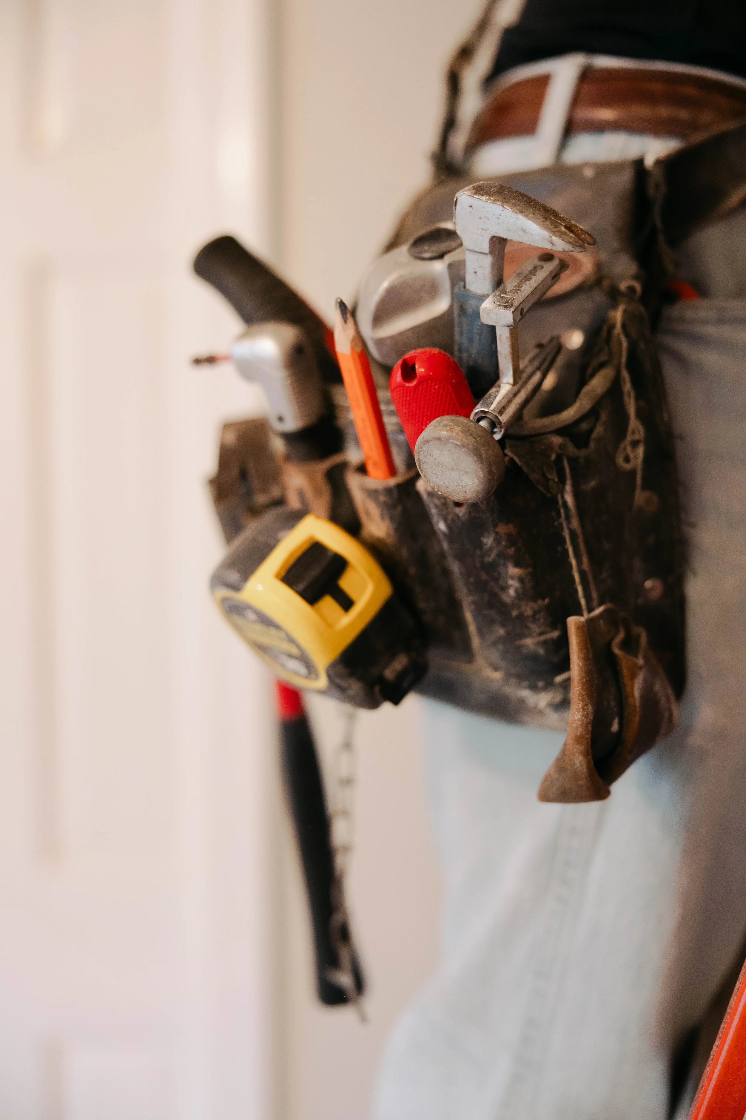 A close-up of a well-used leather tool belt with various tools, including a tape measure, screwdriver, and wrench, hanging on a wall.