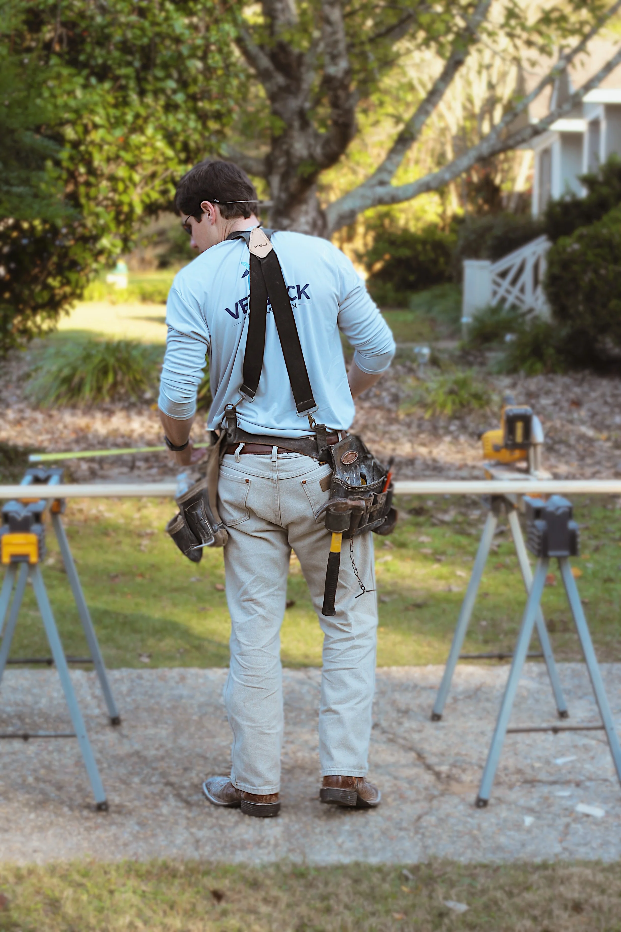 A worker standing outdoors, working on a construction or carpentry project, with a tool belt and tools, surrounded by trees and greenery.