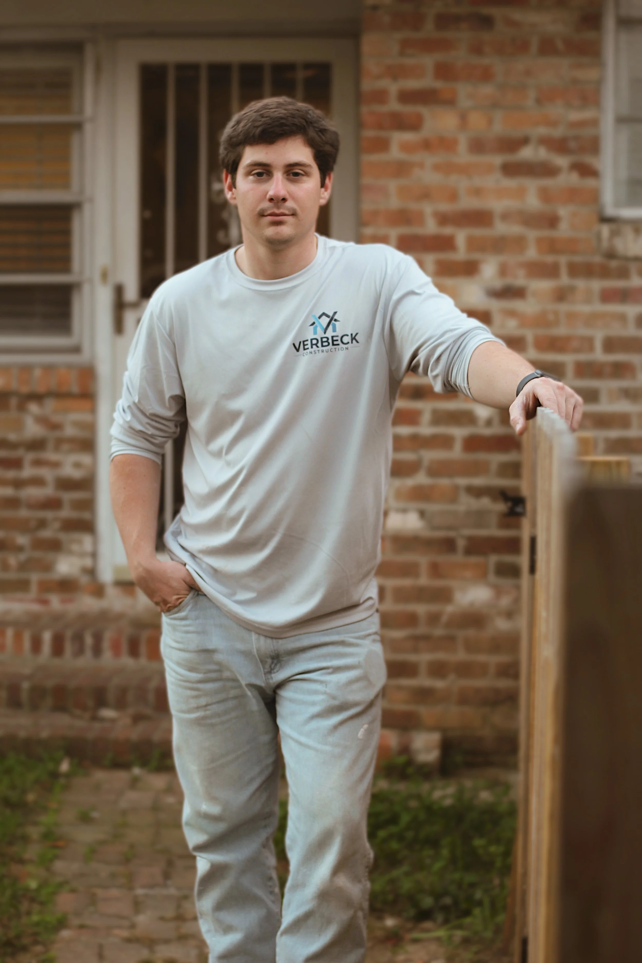 A young man leaning on a wooden fence outside a brick house, wearing a white long-sleeve shirt with a construction company logo and light-colored jeans.