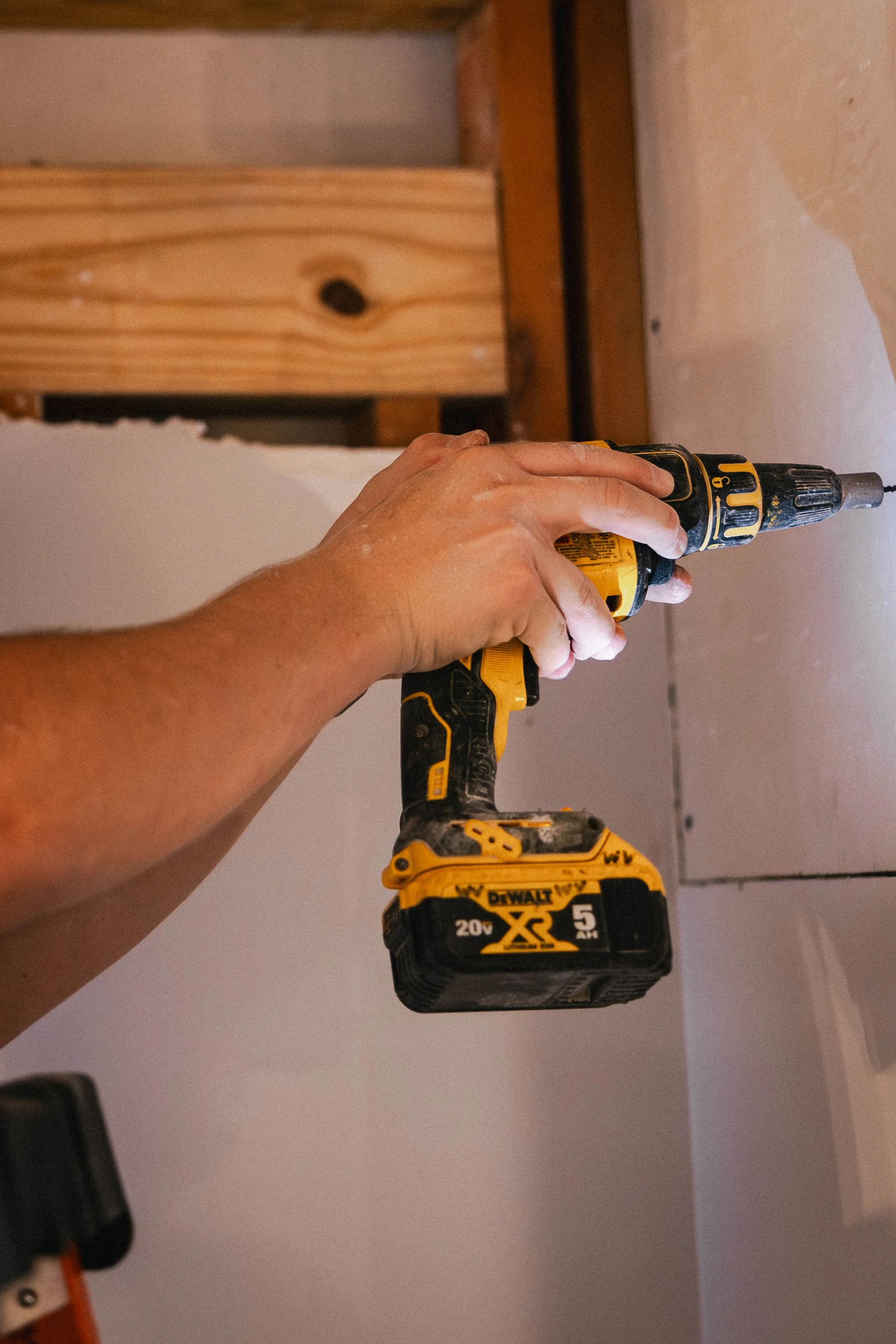 Person using a yellow cordless drill to attach drywall to a wall during construction.
