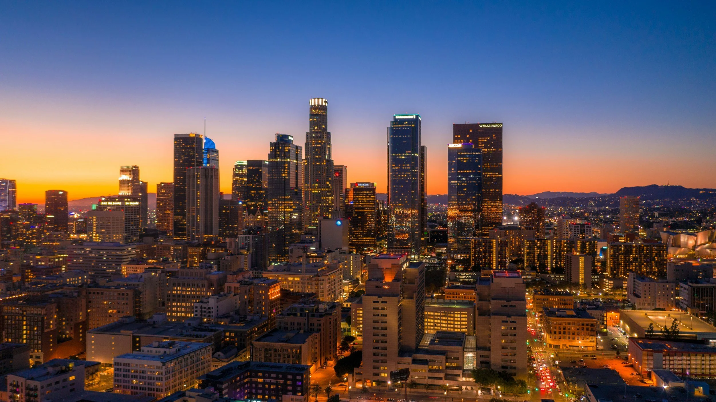 City skyline at sunset with tall skyscrapers illuminated in downtown Los Angeles, California, with mountains in the background.