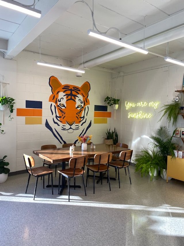 Indoor space with a wall mural of a tiger's face, potted plants, a neon sign that says 'You are my sunshine,' and a wooden dining table with six chairs.
