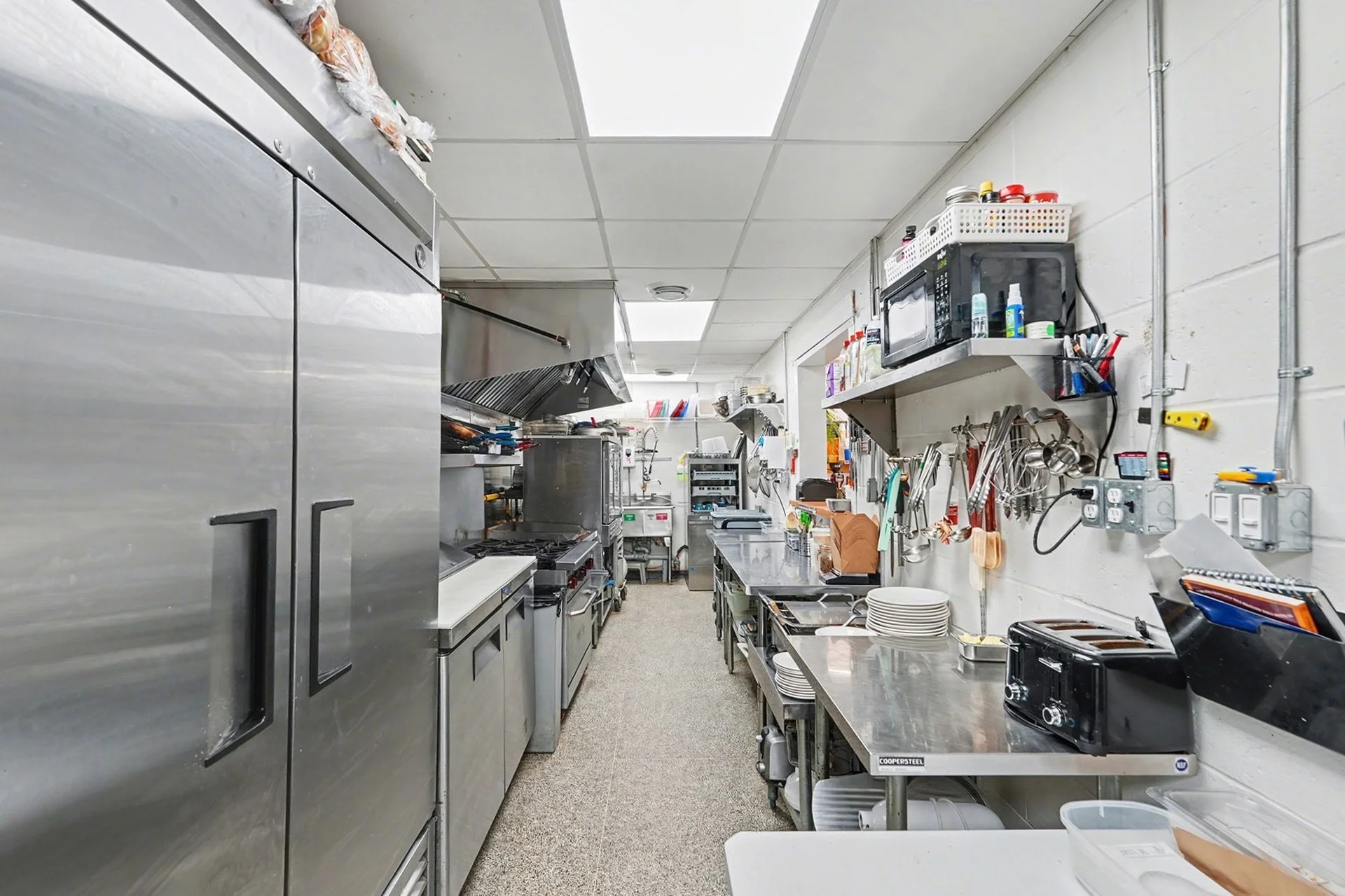 Commercial kitchen with stainless steel appliances, counters, and cooking equipment along both sides, with utensils, plates, and appliances organized on the wall and counters.