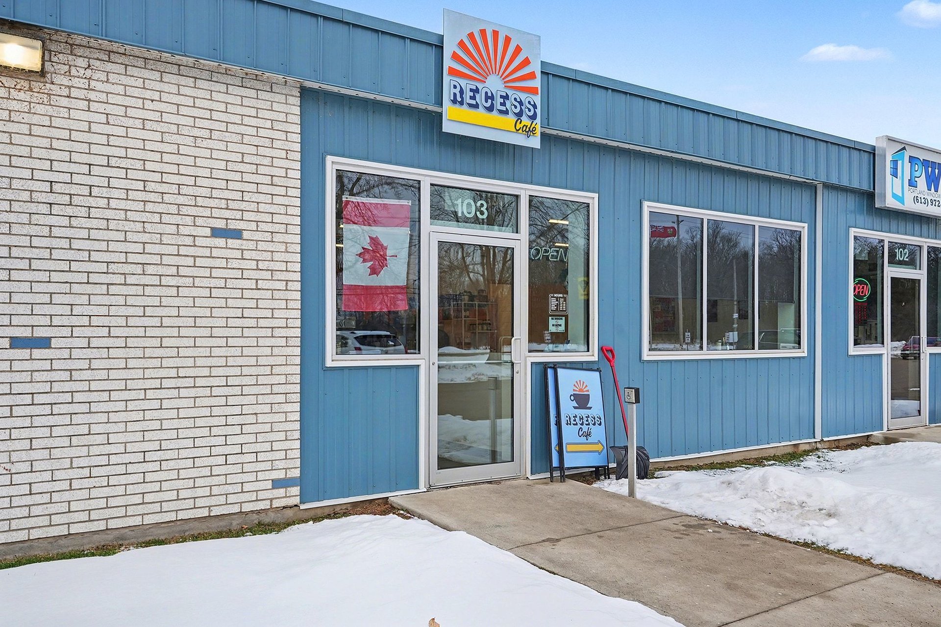 Exterior view of Recess Cafe, a small storefront with blue siding and white brick, with a large window displaying a Canadian flag. There is a sidewalk and snow on the ground, with a sign and a shovel outside indicating the cafe's entrance.