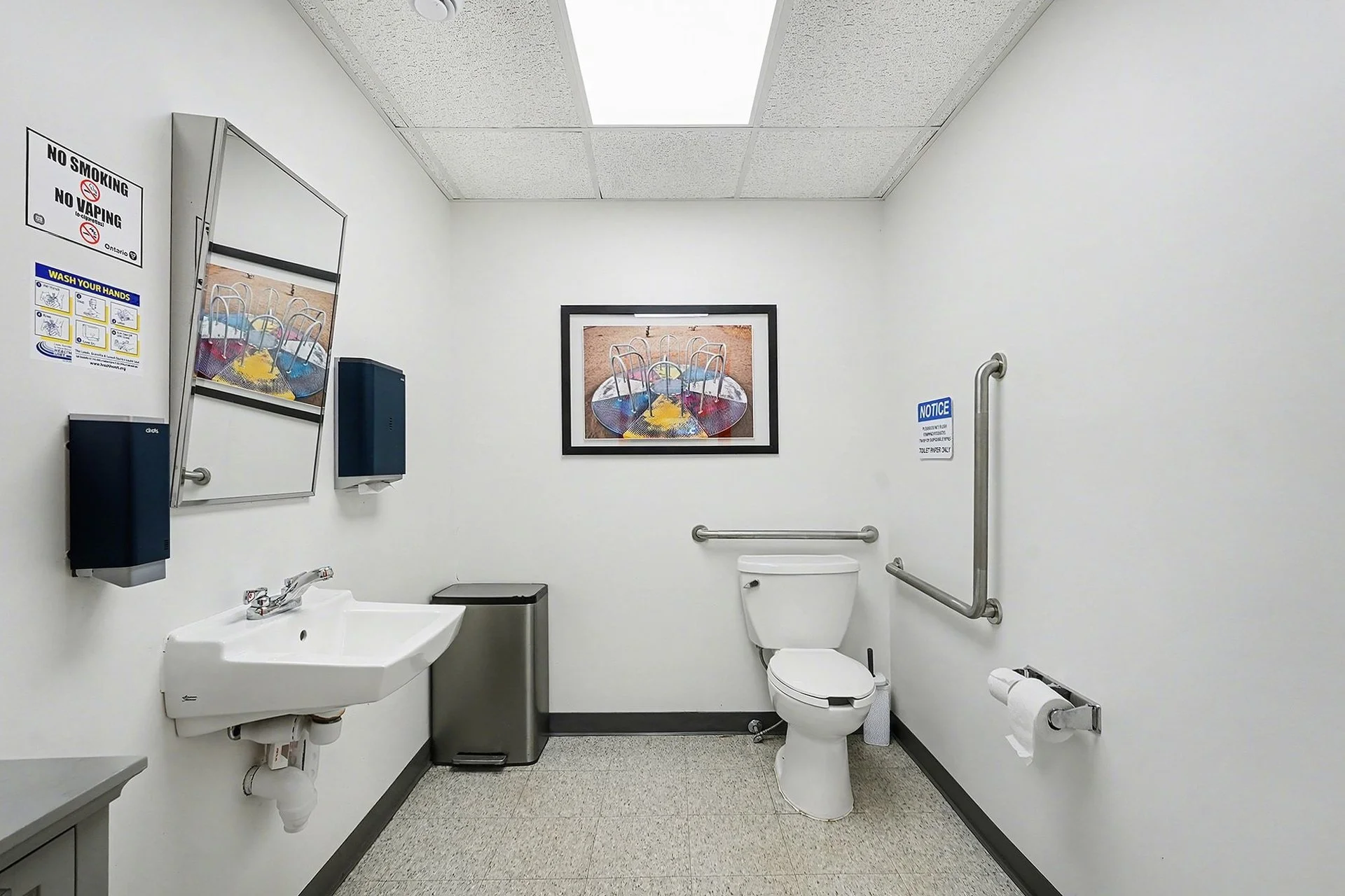 A clean, white bathroom with a toilet, sink, paper towel dispenser, and framed painting of chairs around a table on the back wall.