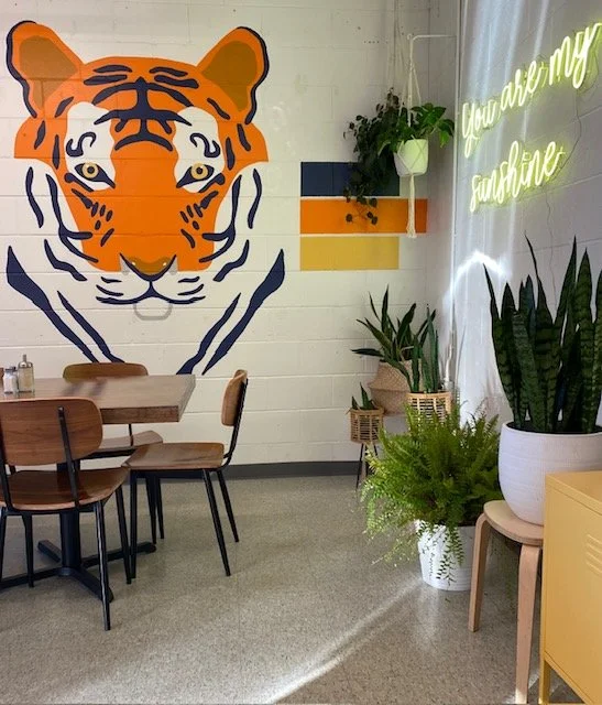 Interior of a cafe or restaurant with a tiger mural on the wall, a neon sign that reads "You are my sunshine," and various potted plants.