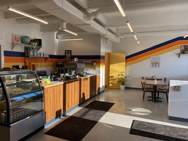 Coffee shop interior with a display case, counter, and seating area, featuring white walls with colorful stripe accents, and a yellow door.