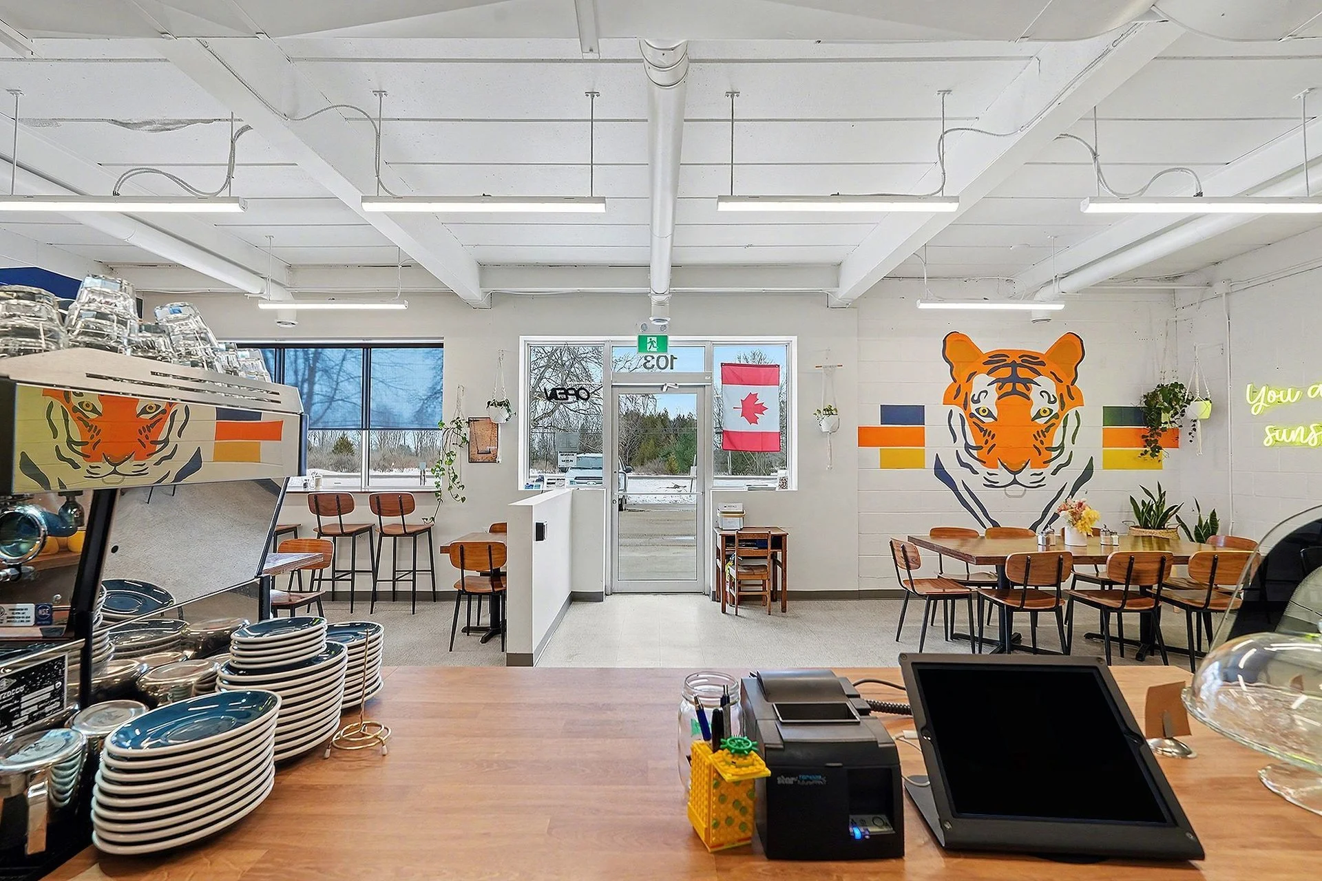 Interior of a modern restaurant or café with wooden tables and chairs, a tiger mural on the wall, potted plants, neon sign, and large windows showing an outdoor view.