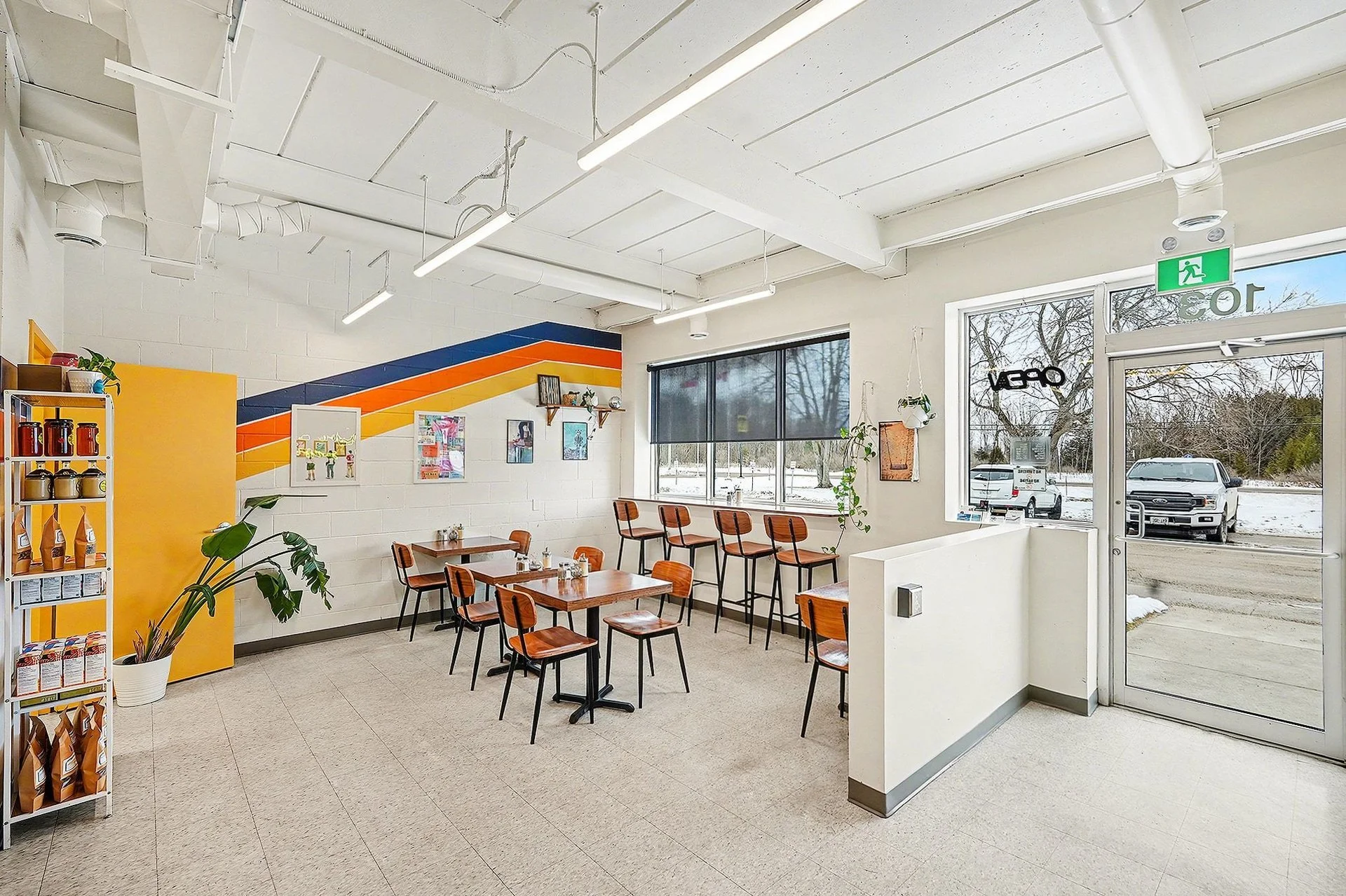 Interior of a casual cafe with wooden tables and chairs, large windows, colorful wall art, indoor plants, and an exit door to the outside parking lot covered in snow.