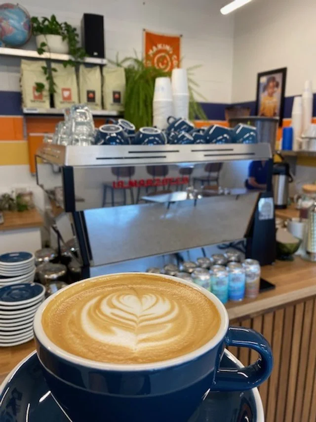 A blue mug of latte with latte art on top in a coffee shop. Behind it is an espresso machine with cups arranged on top, and shelves with various items like plants, glasses, and decorative pieces.