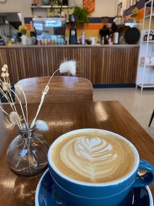 A cup of latte with latte art in a blue mug, on a wooden table next to a small decorative vase with dried flowers, inside a cozy coffee shop with a counter and equipment in the background.