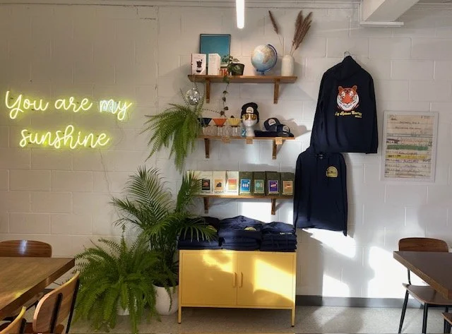 Decorative wall with a yellow neon sign saying 'You are my sunshine,' two wooden shelves holding books, a globe, plants, and decorative items. The wall has hanging clothes, including a hoodie with a tiger graphic. There are plants on top of a yellow 