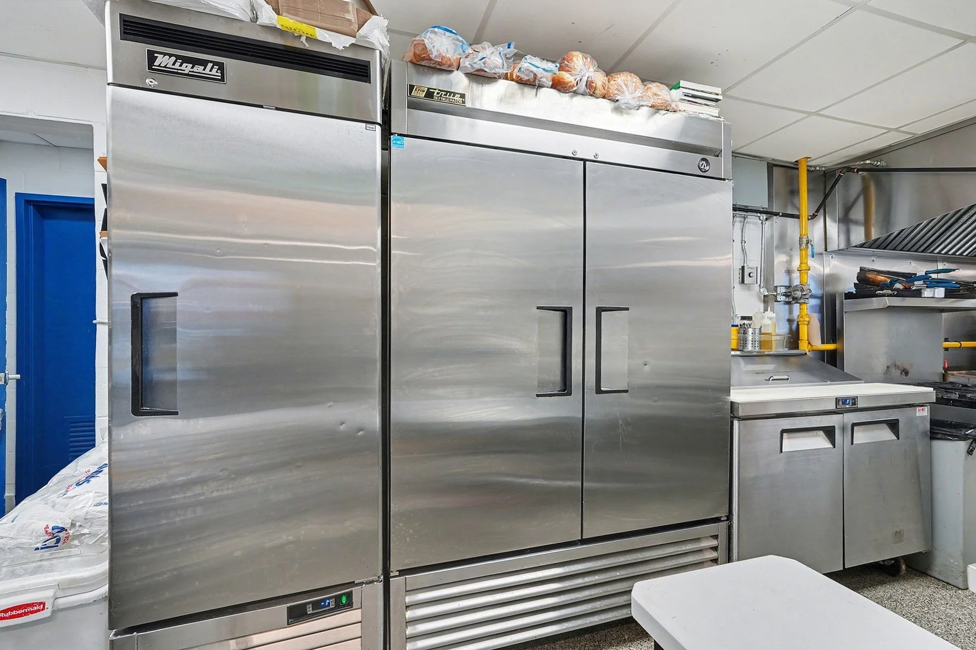 Commercial kitchen with stainless steel appliances, including a large refrigerator and freezer, with plastic bags of vegetables on top, yellow gas pipes, and kitchen tools on the countertop.