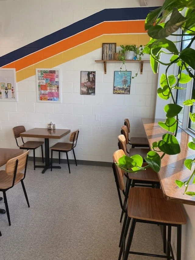 Corner of a cozy cafe or dining area with wooden tables and chairs, a white brick wall decorated with paintings and a small wooden shelf holding books and plants, and a large window with hanging green plants.