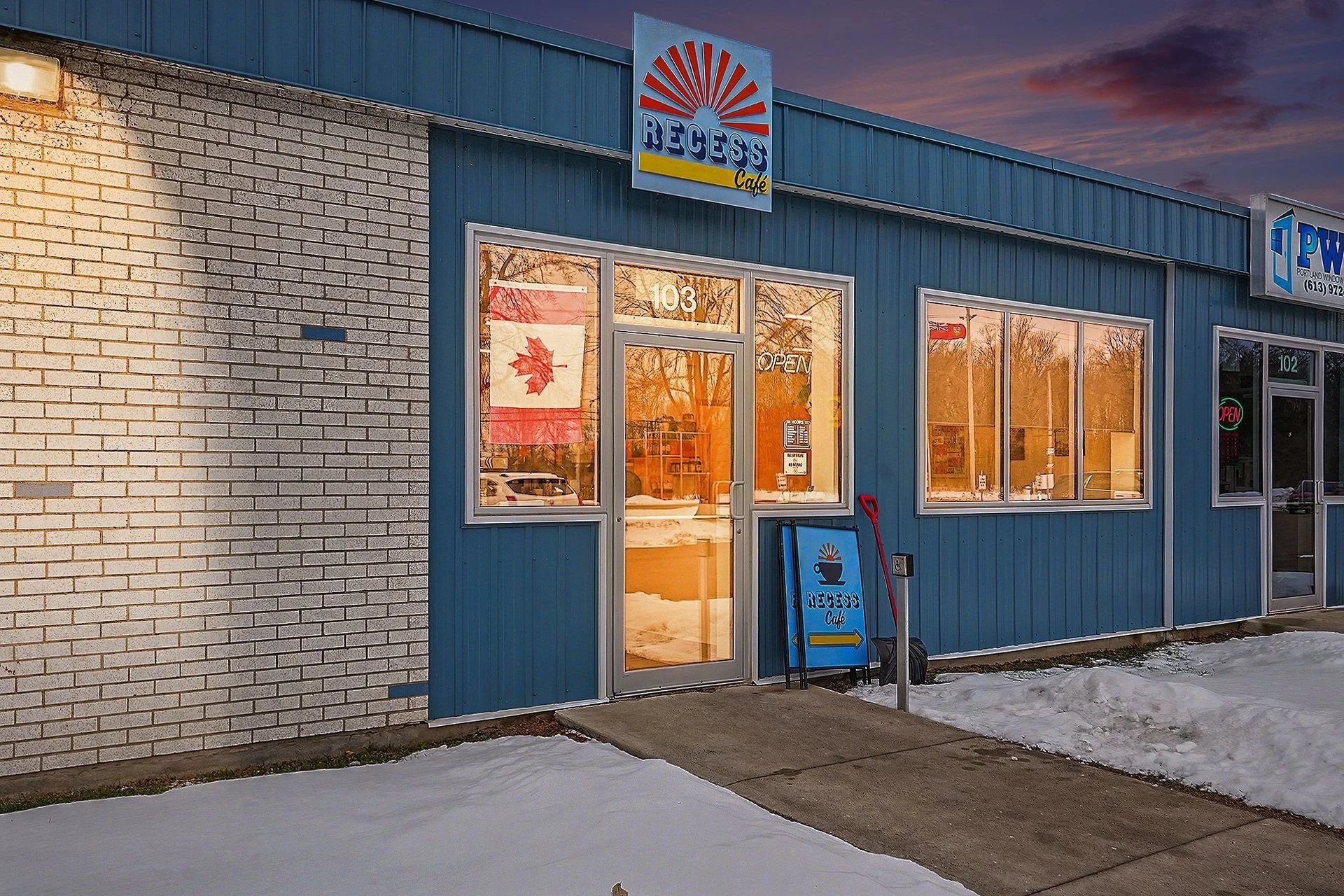 Exterior of Recess Café with a blue facade, large windows, and a sign above the door. Snow on the ground, a Canadian flag visible inside the window, and a sidewalk leading to the entrance during dusk.