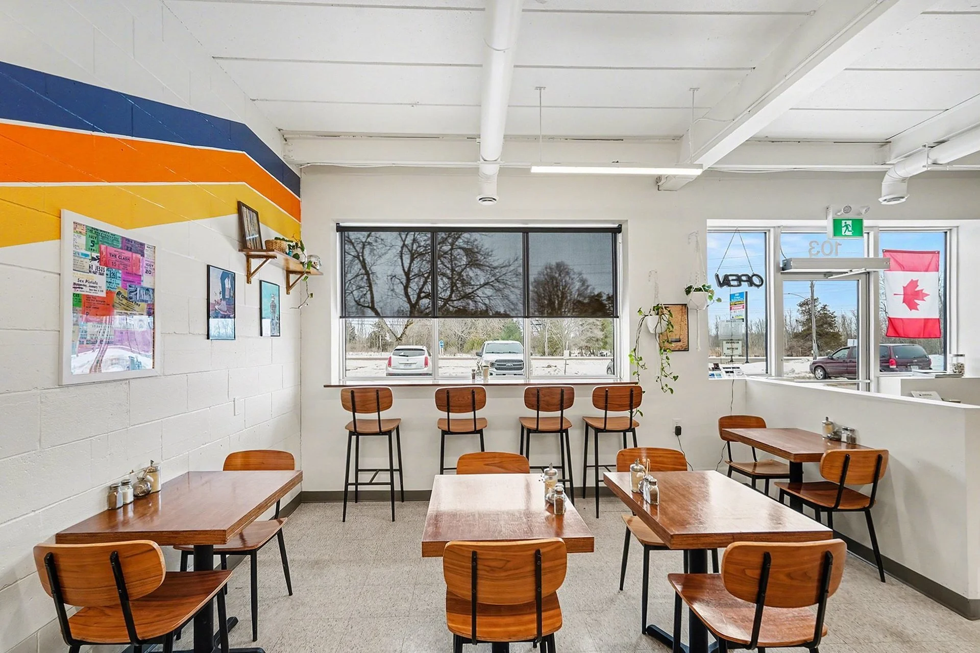 Empty café with wooden tables and chairs, large front window, Canadian flag, and interior wall with colorful stripe pattern.