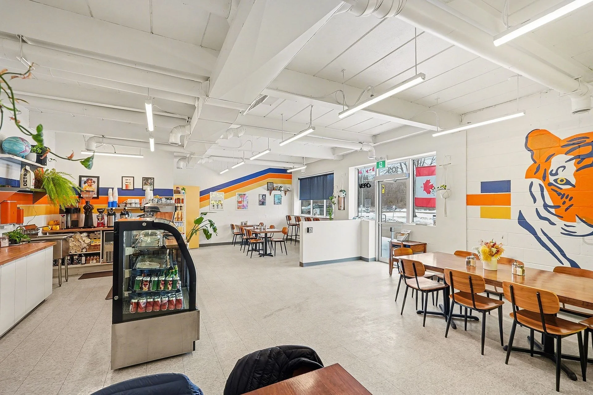 Interior of a bright cafe with white walls, colorful striped wall art, wooden tables and chairs, a small refrigerator with drinks, and a coffee station. Large windows with a Canadian flag and snow outside.