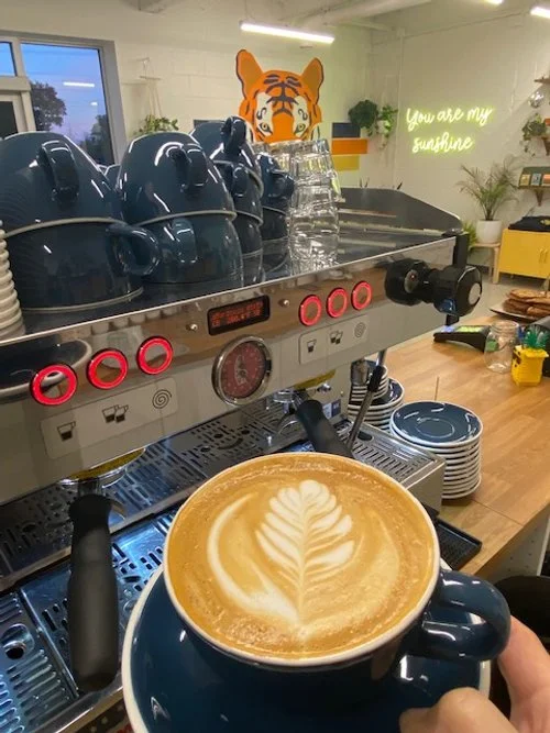 A cup of latte with leaf foam art in front of a mocha pot espresso machine, with blue teapots and a tiger head mask on top of the machine, in a cozy café setting.