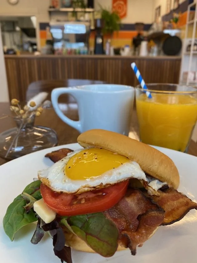 Plate with bacon, fried egg, tomato, cheese, and lettuce, alongside a glass of orange juice and a white mug on a restaurant table.