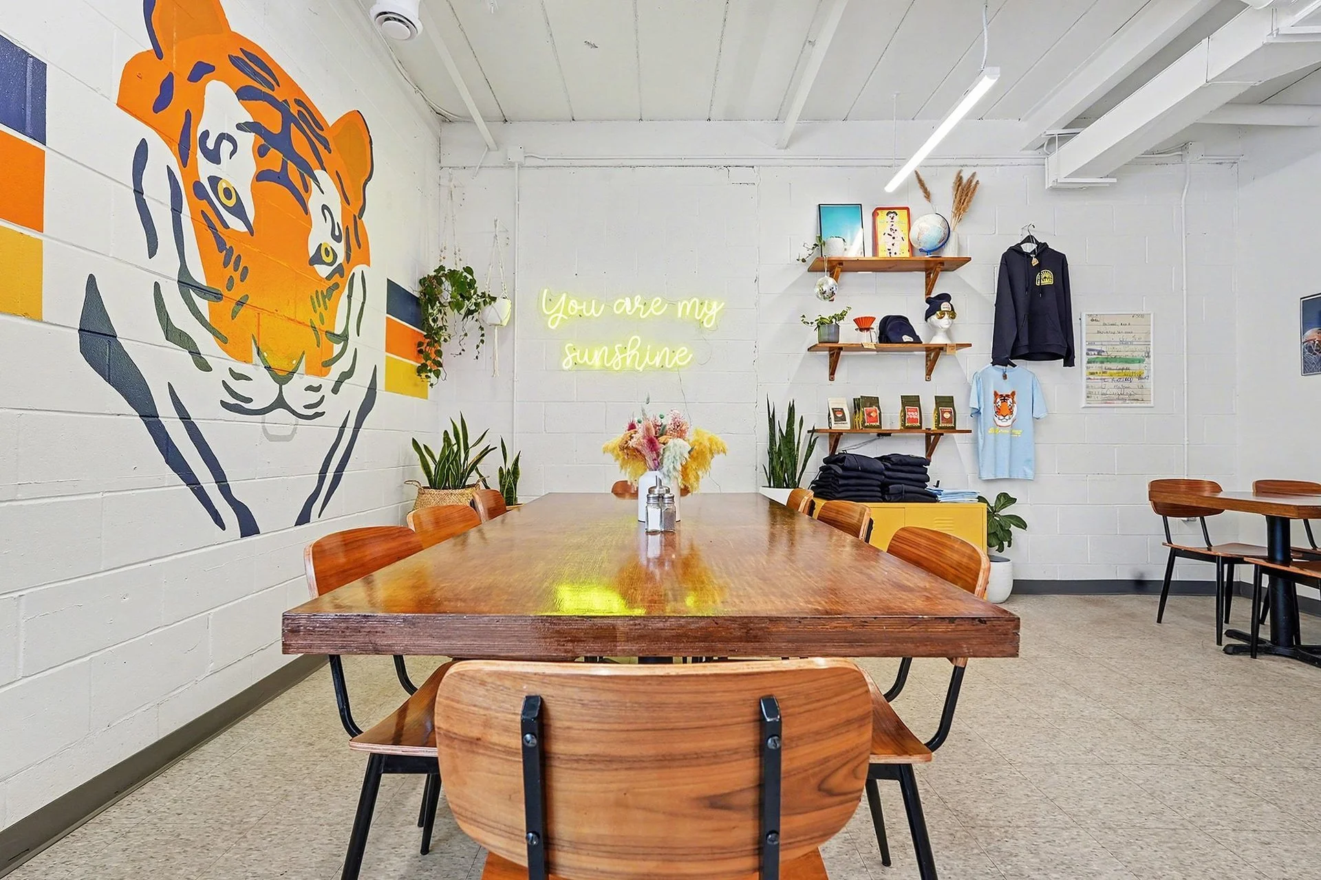 Interior of a cafe or shop with a wooden table, surrounded by wooden chairs, with a large tiger mural on the wall. The wall has a neon sign that reads 'You are my sunshine', shelves with books, plants, and clothing, and various decorative items.