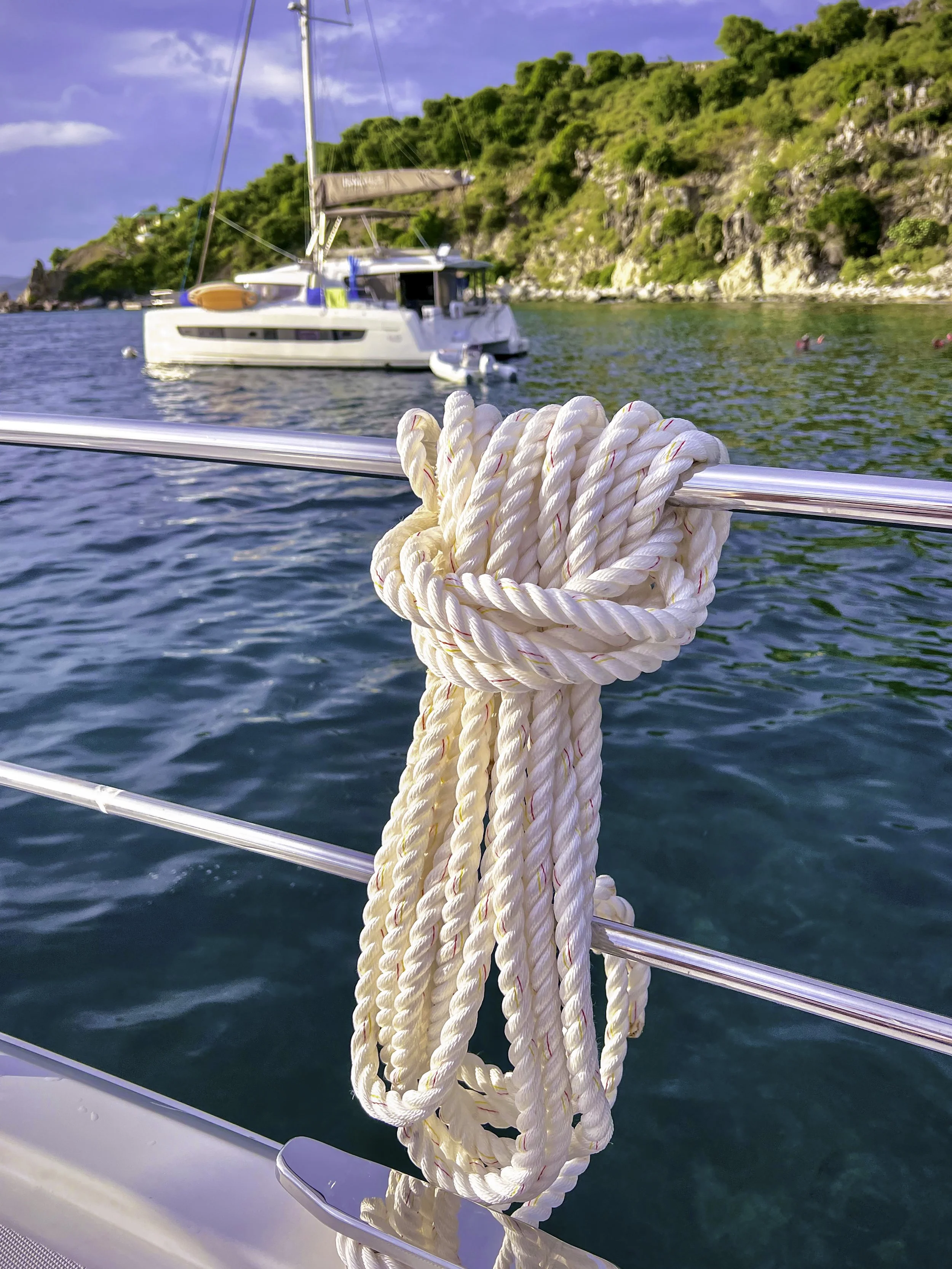 View of a white rope tied to a metal railing on a boat, with a green hill, a sailboat, and a blue sky with some clouds in the background.