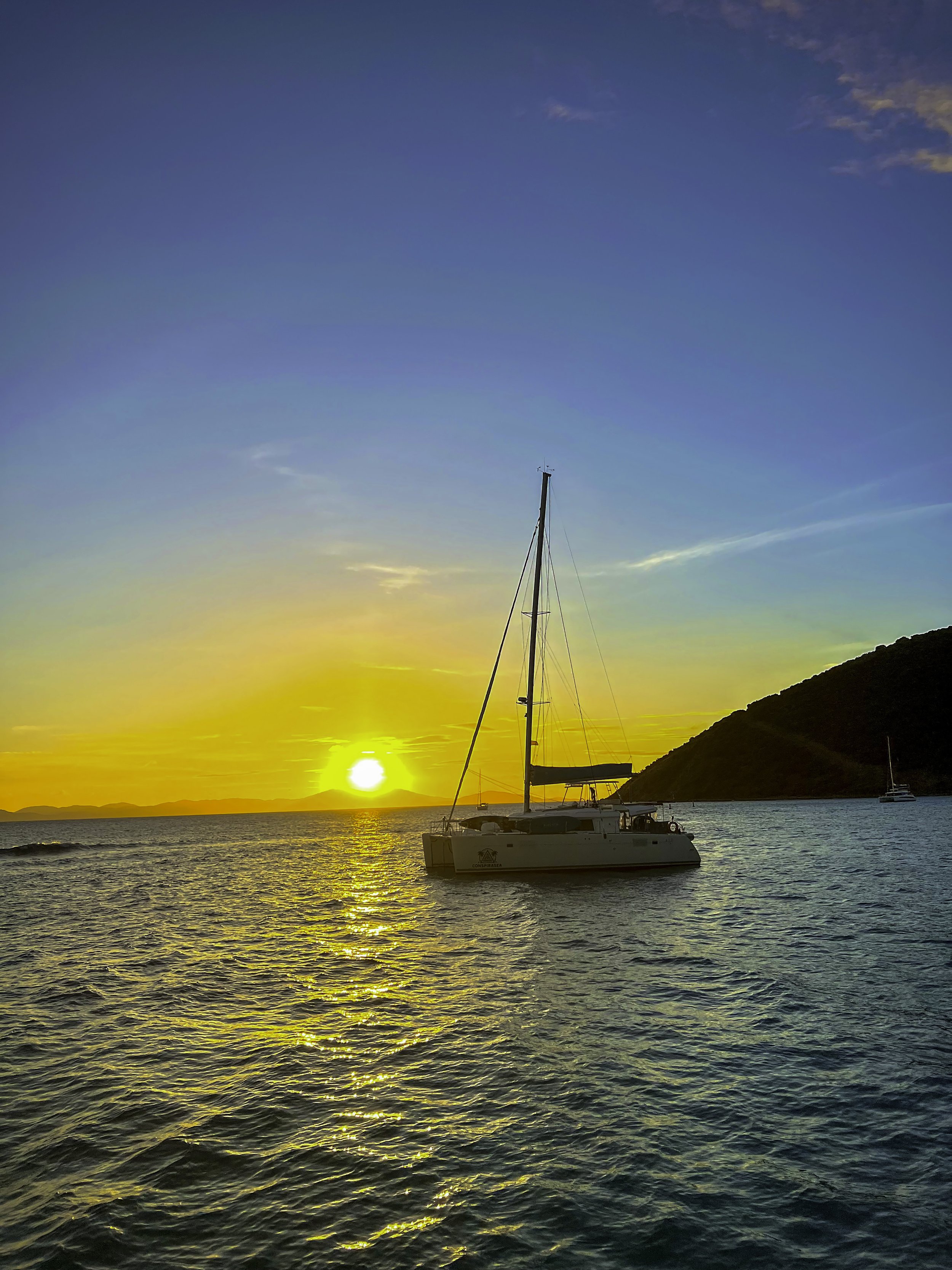 Sunset over the ocean with a sailboat anchored near the coast, a hill in the background, and a partly cloudy sky.