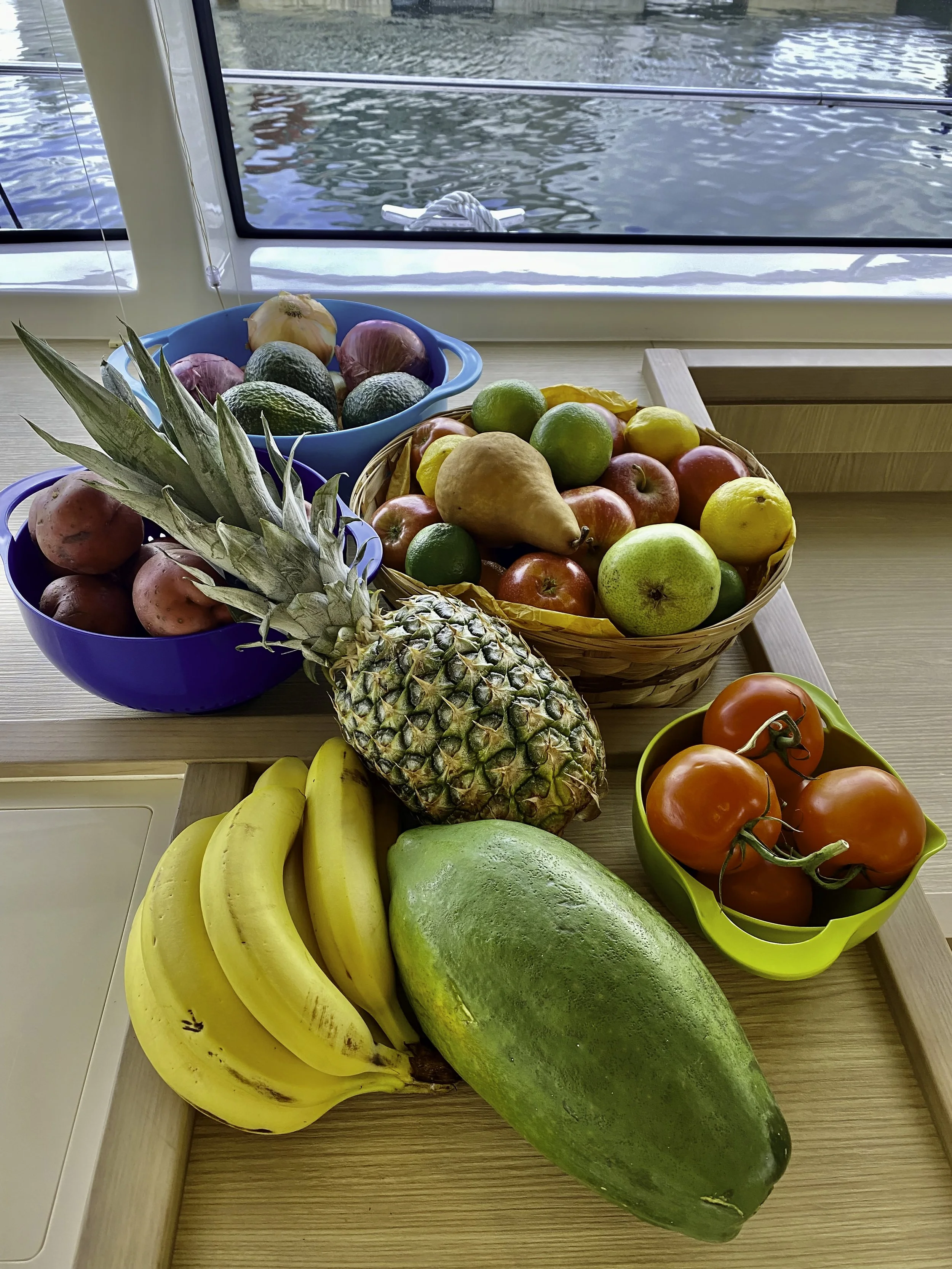 A variety of fresh fruits on a wooden table near a window, including bananas, a watermelon, pineapple, tomatoes, an apple, pear, lime, avocado, and other fruits.