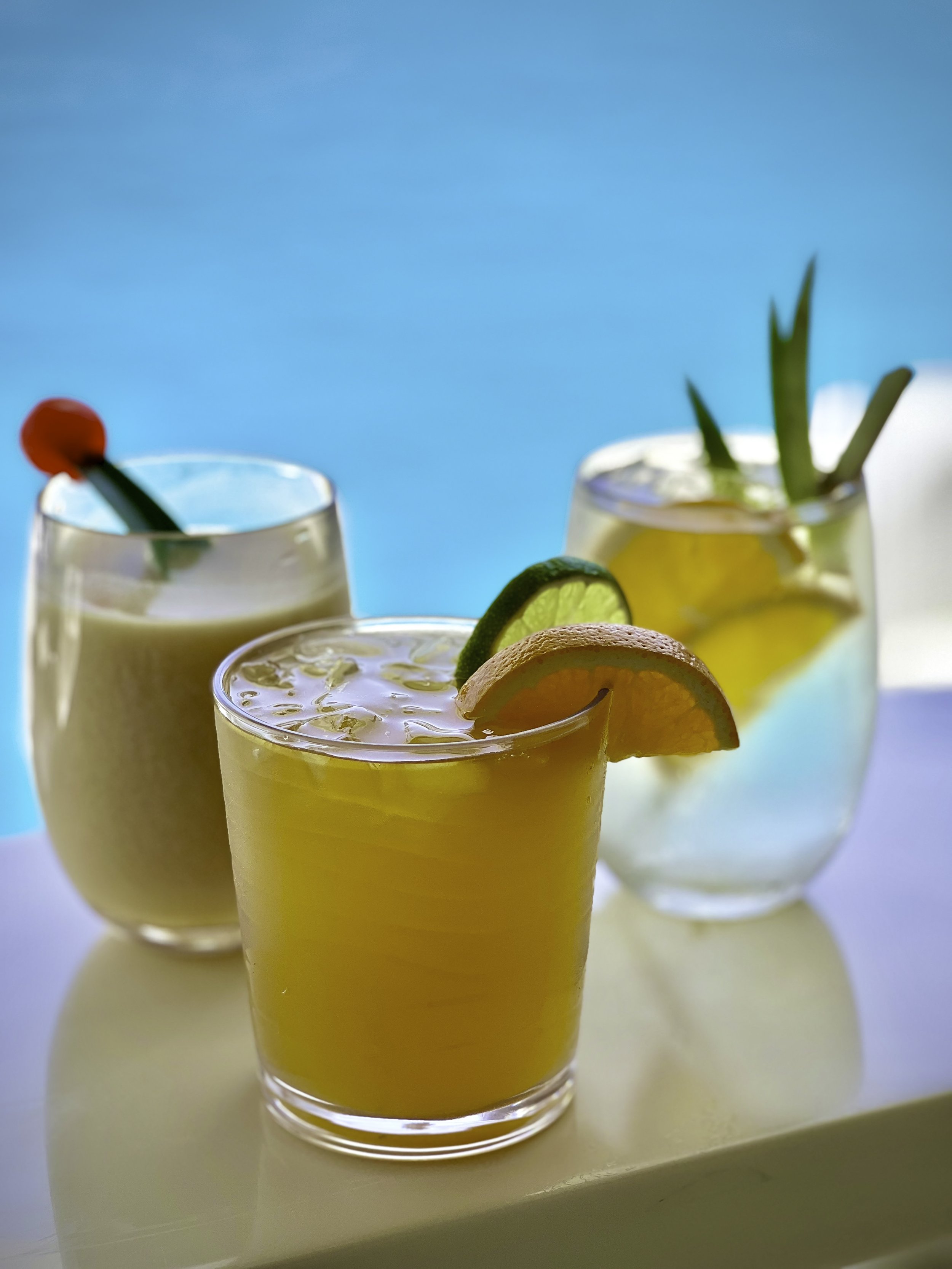 Three colorful cocktails with garnishes on a white surface against a bright blue background.