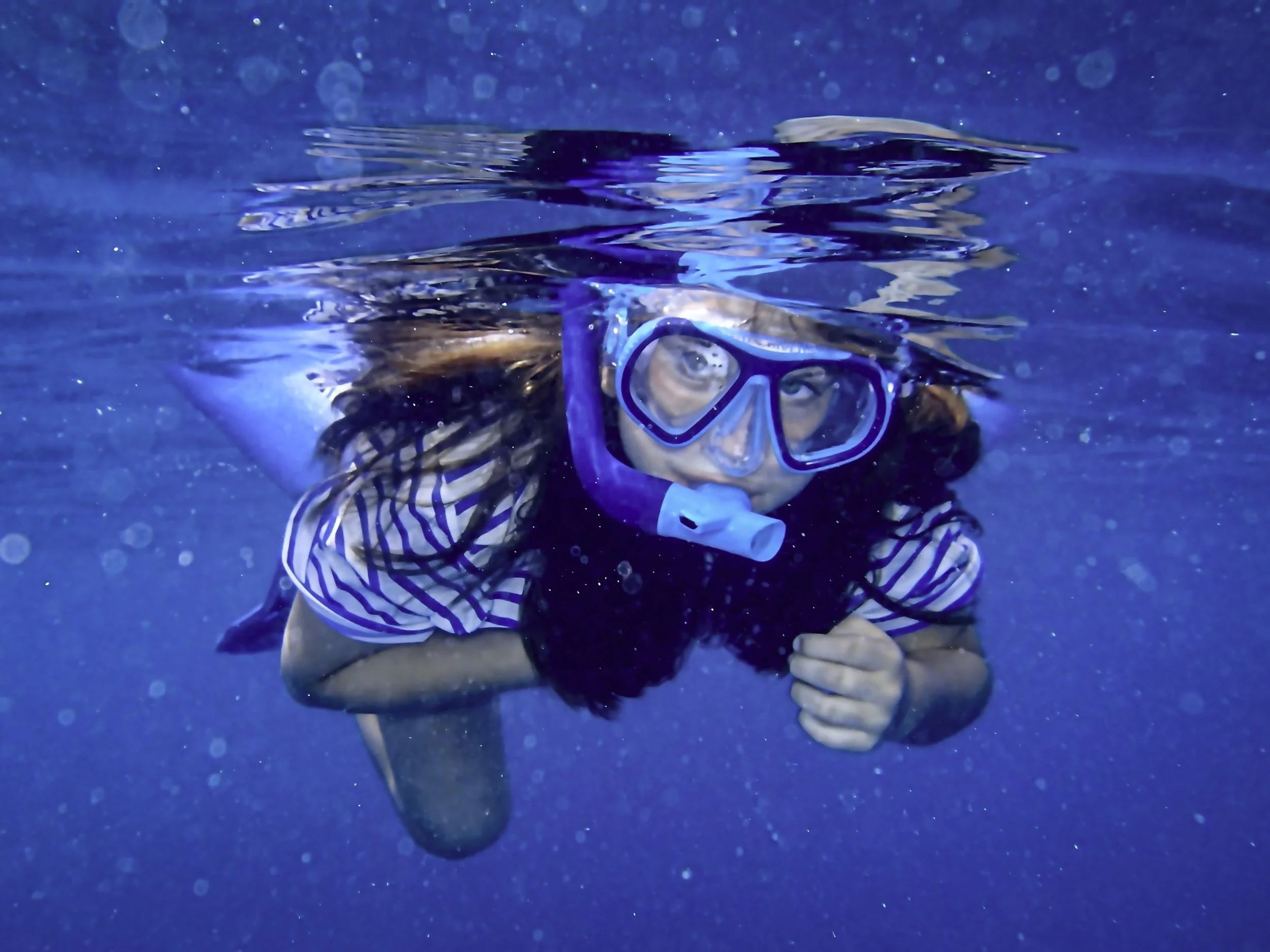 A woman with long hair snorkeling underwater, wearing a blue mask and snorkel, and a striped shirt.