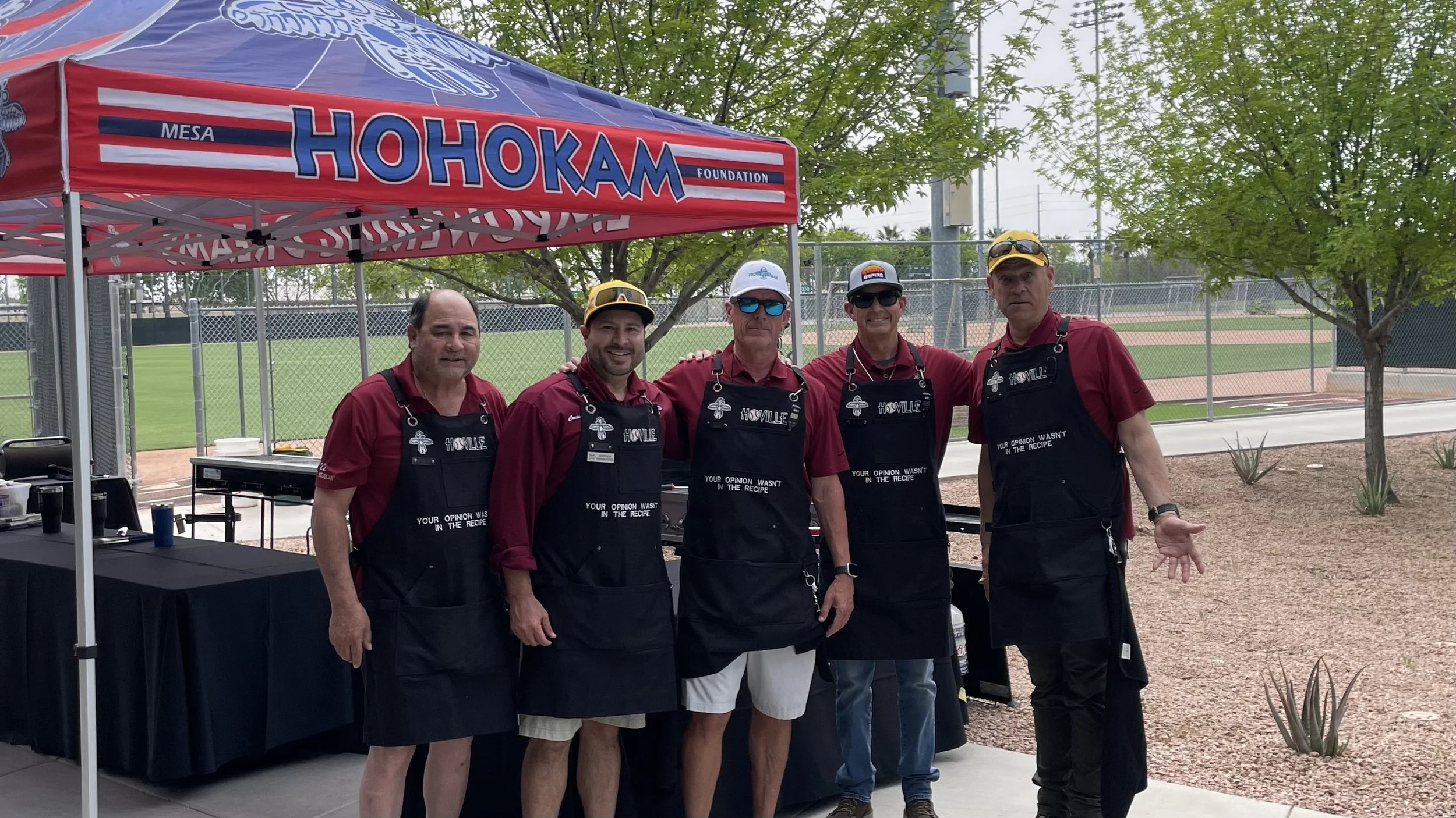 Five men standing side by side outdoors at a baseball field, wearing matching maroon shirts and black aprons with text, under a red canopy tent with the words 'HOHKAM Foundation' and 'Mesa' visible, surrounded by trees and greenery.