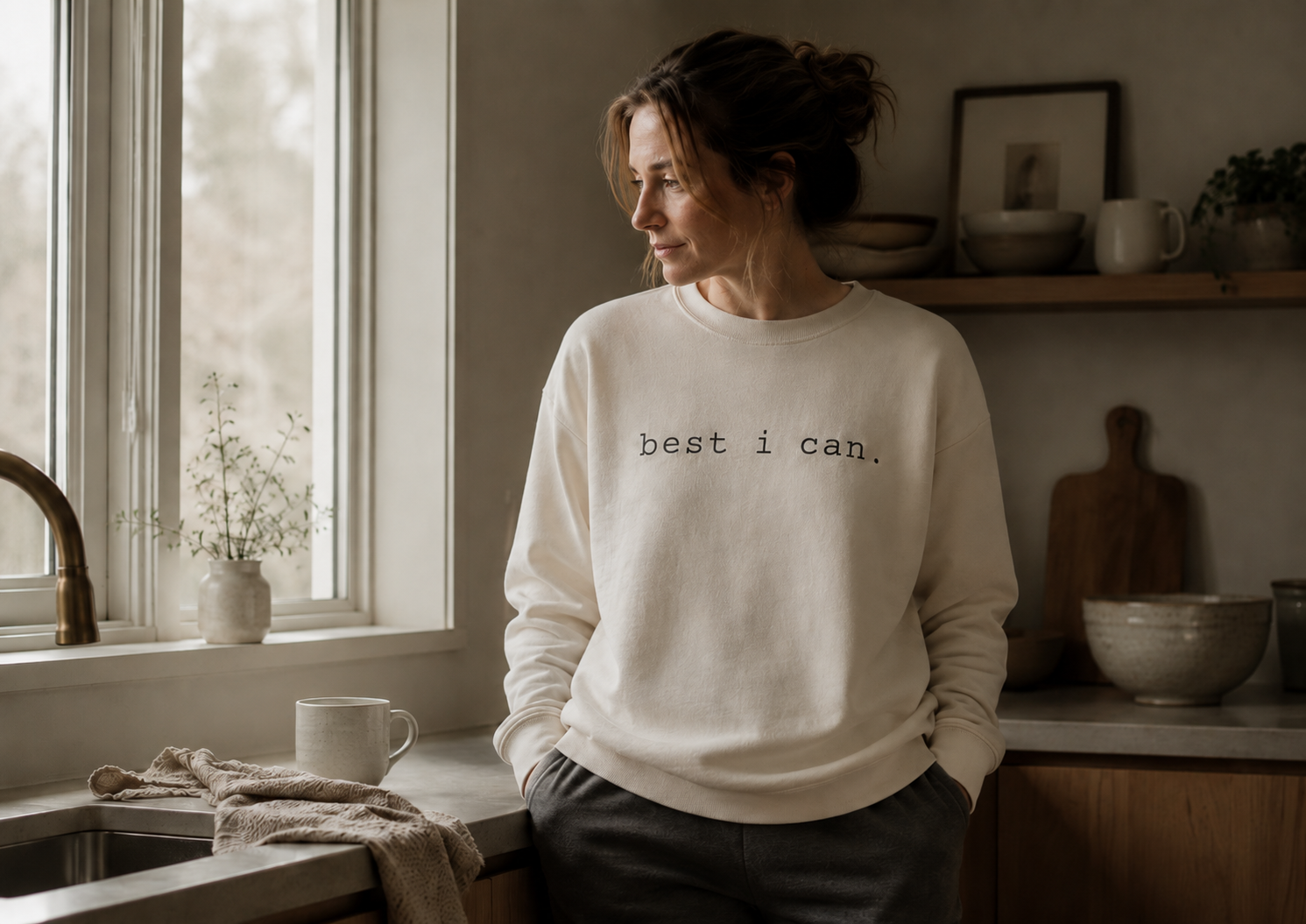 person wearing a cream best i can sweatshirt standing in a softly lit kitchen beside a window