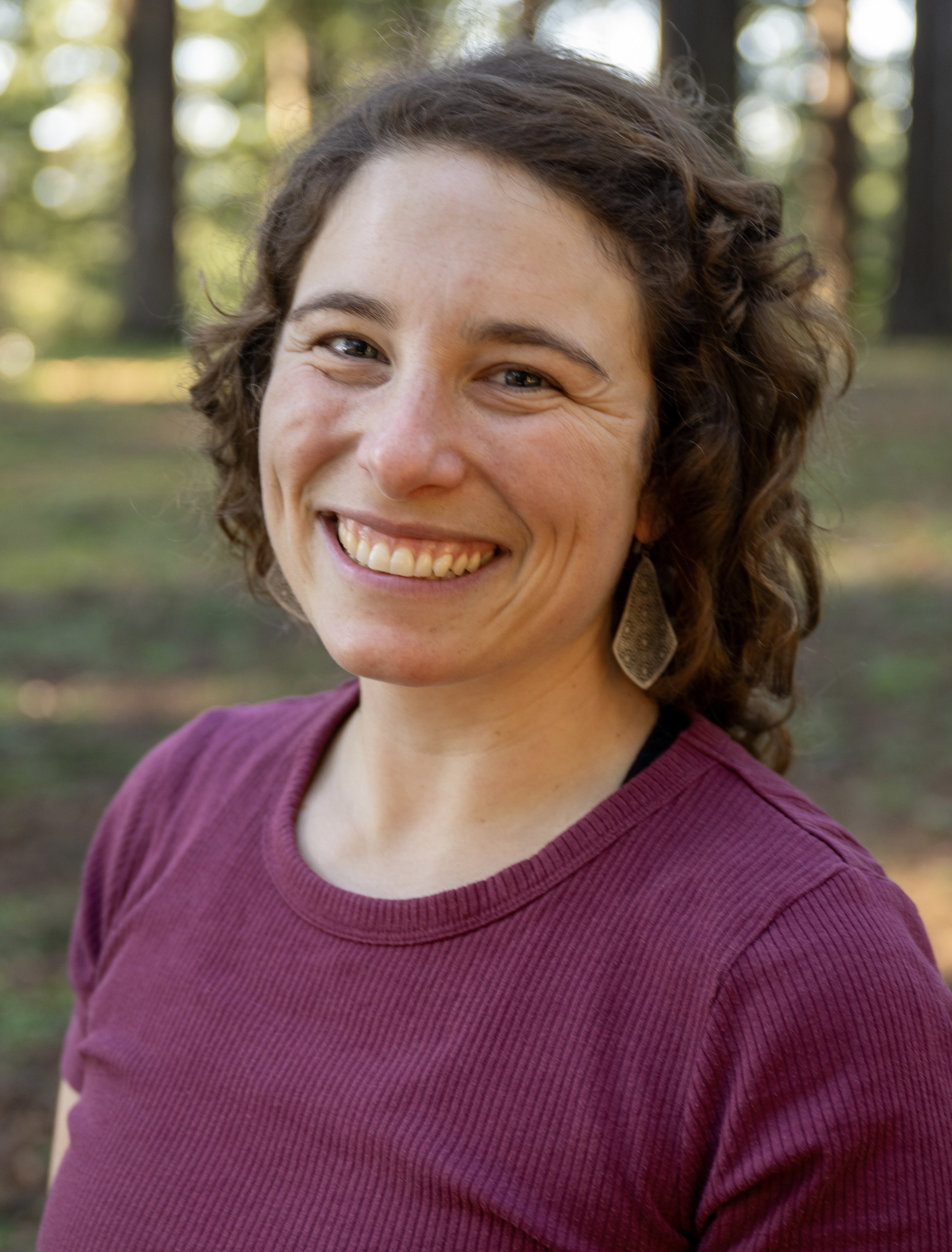 A woman with curly brown hair smiling, wearing a burgundy top and patterned earrings, standing outdoors in a wooded area with trees and sunlight in the background.